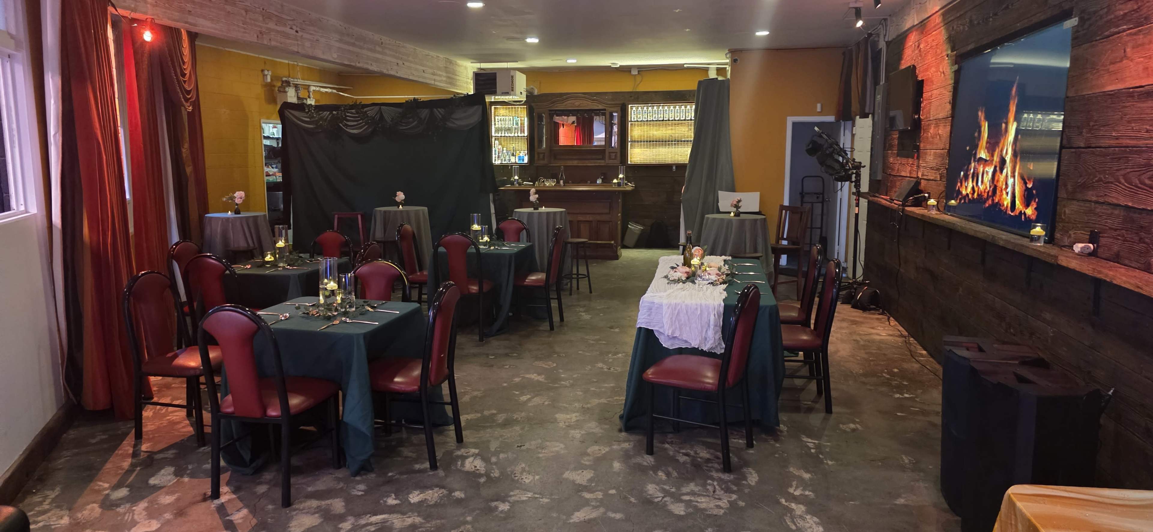 The image shows a cozy dining area with several tables covered in dark green cloths, arranged around a bar and featuring a decorative wall with a fireplace display.