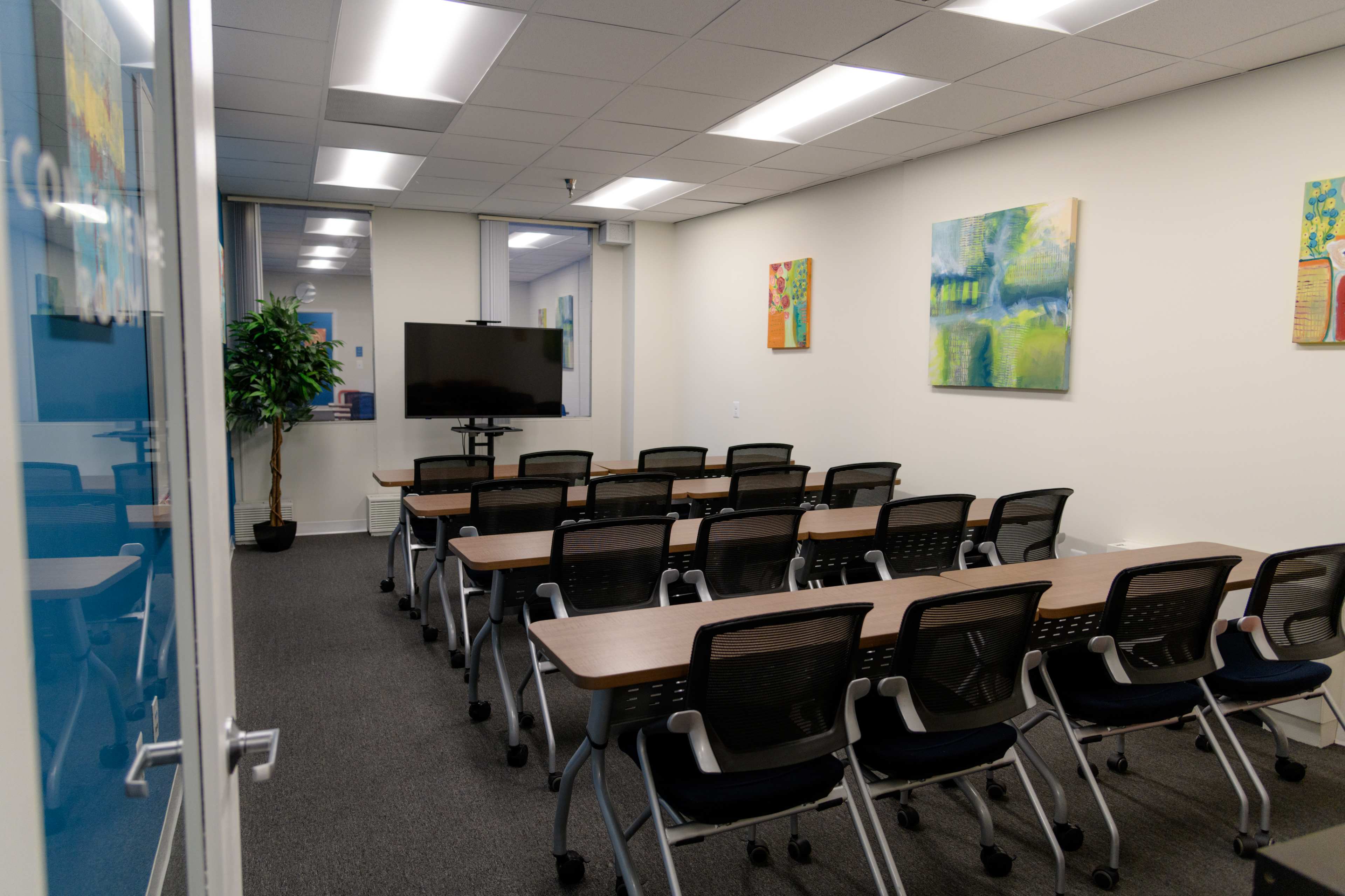 A conference room features several rows of tables and chairs facing a wall-mounted television, with colorful artwork on the walls.