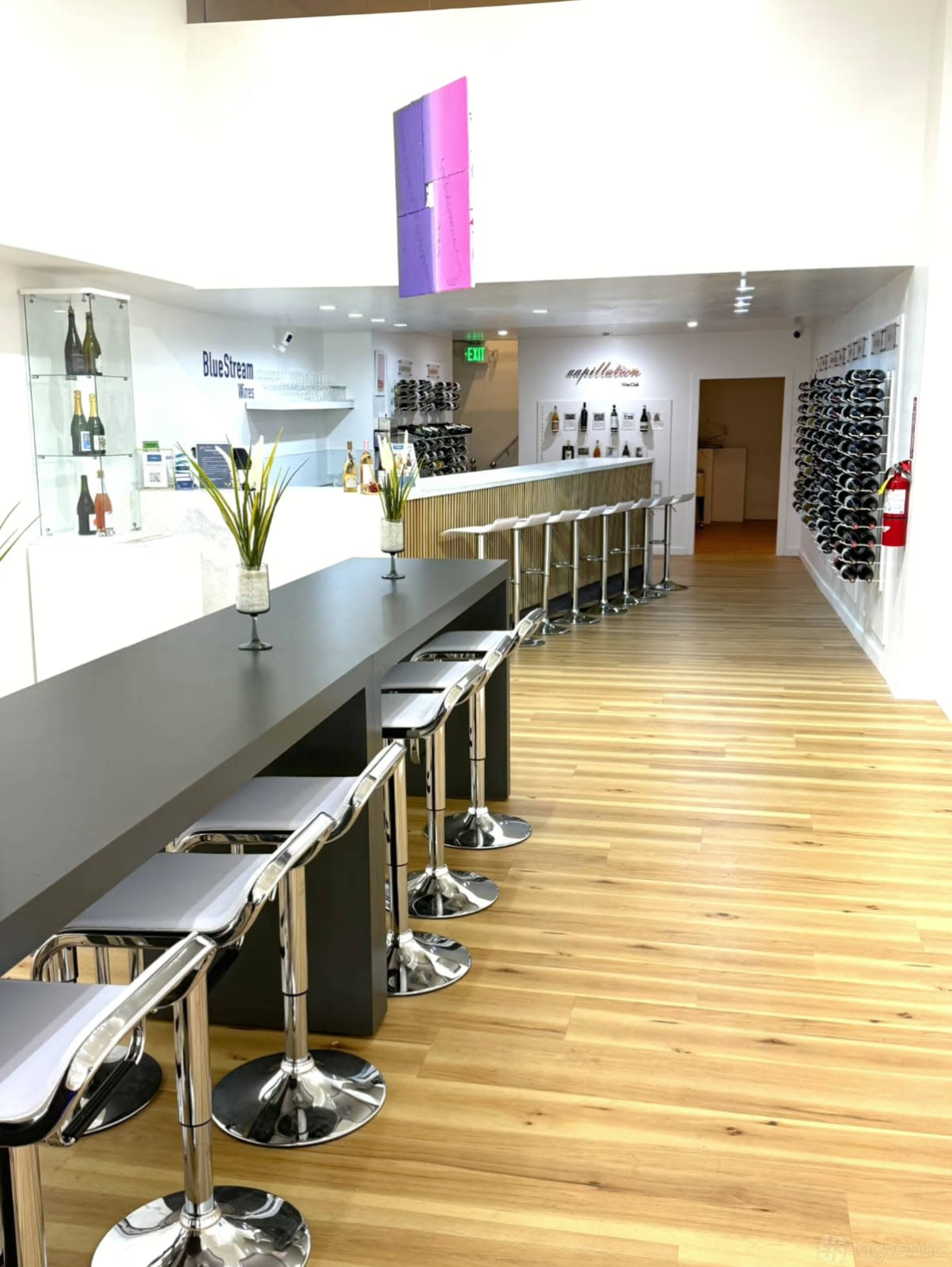 The image shows a modern interior space featuring a long bar with high stools, a display of wine racks on the right, and retail shelves on the left.