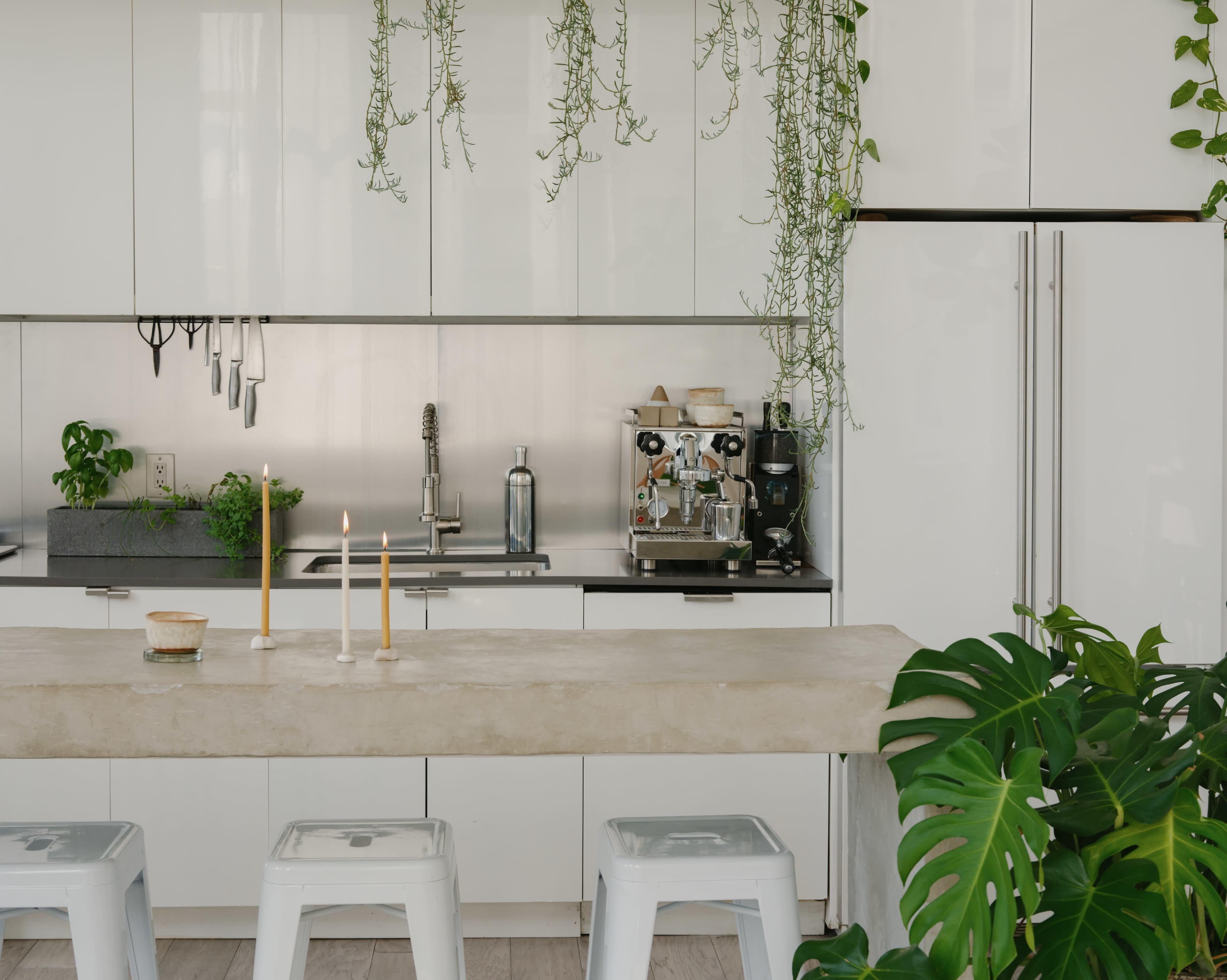 A modern kitchen features a concrete island with two candles, metal stools, and greenery hanging from the ceiling, alongside sleek cabinetry and kitchen appliances.