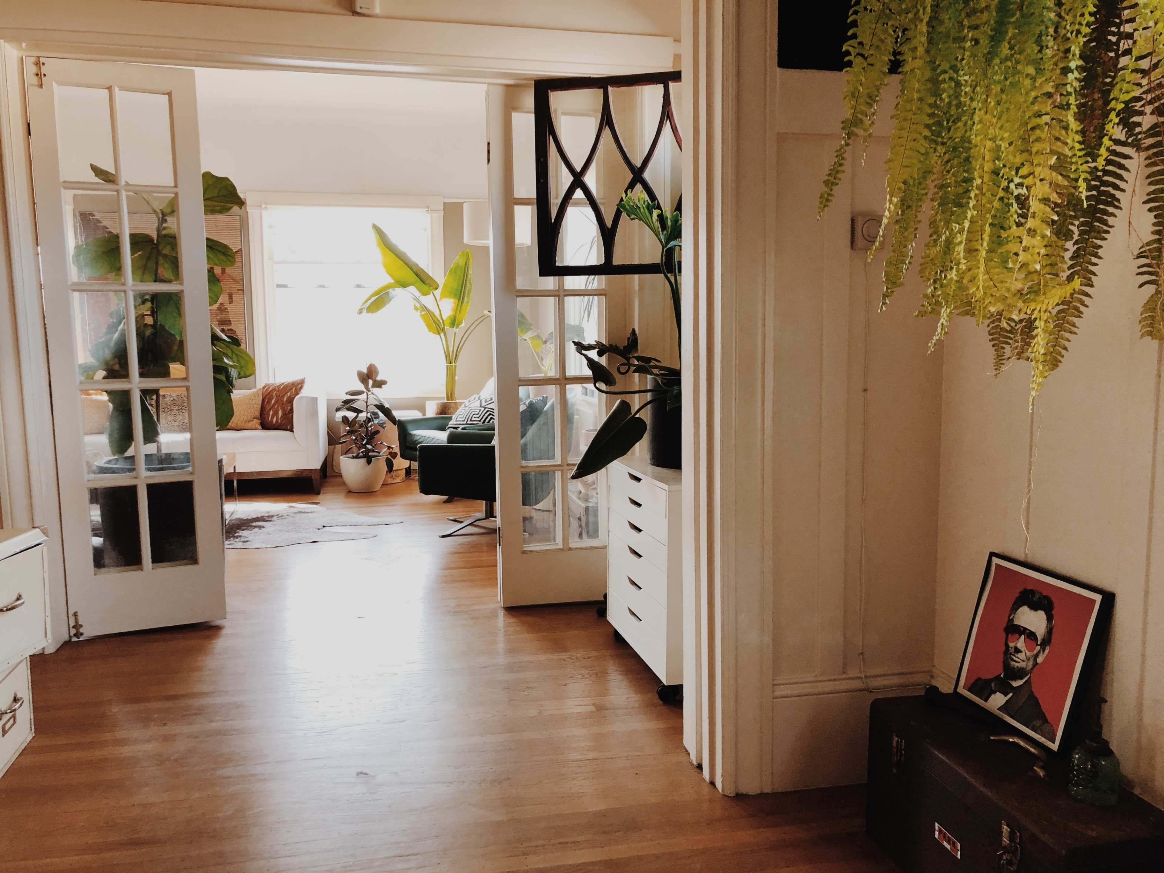 A hallway leads into a bright living space with plants and furniture, framed by glass-paneled doors and decorated with a portrait on a side table.