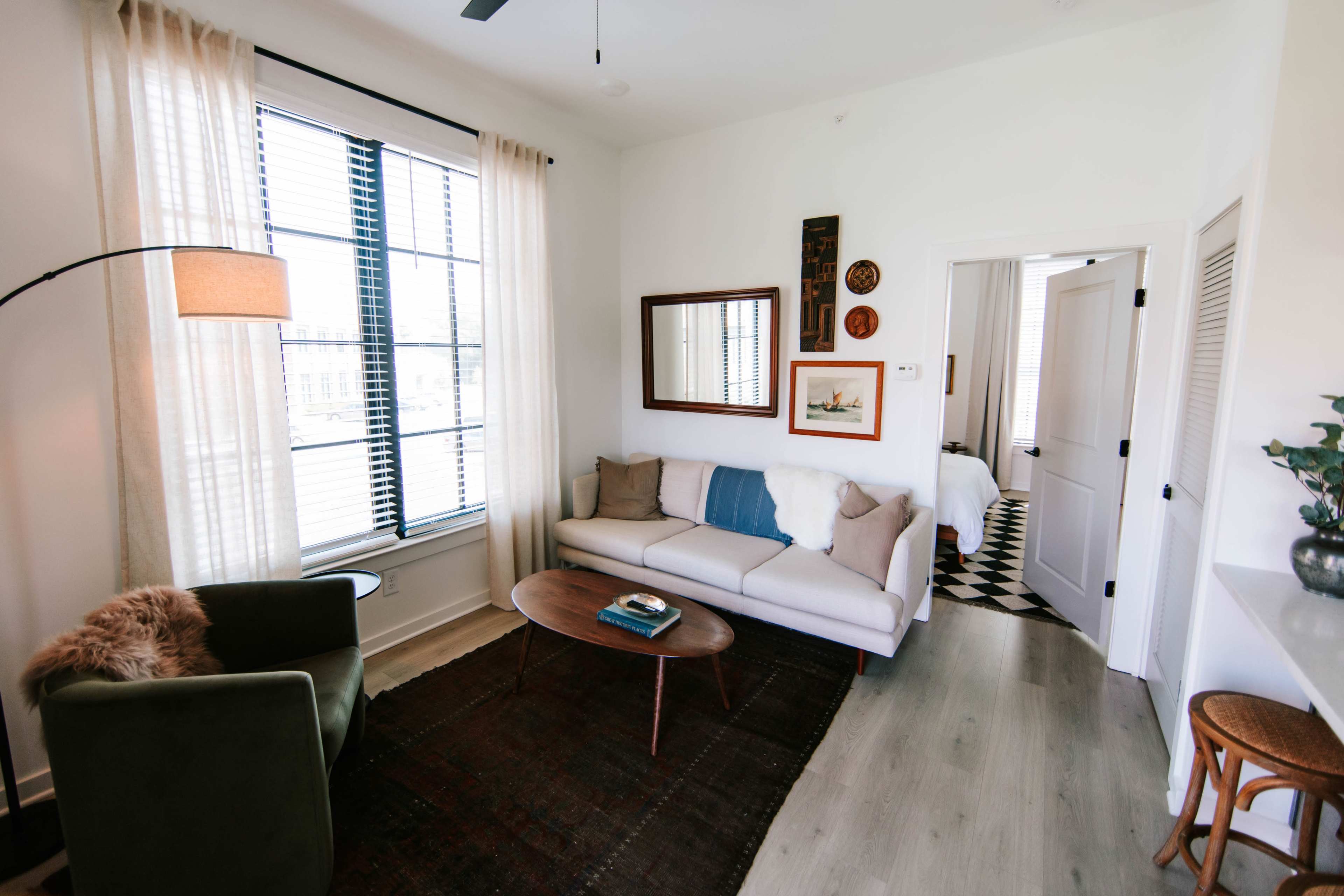 The image shows a cozy living room with a sofa, armchair, coffee table, and a large window with sheer curtains.