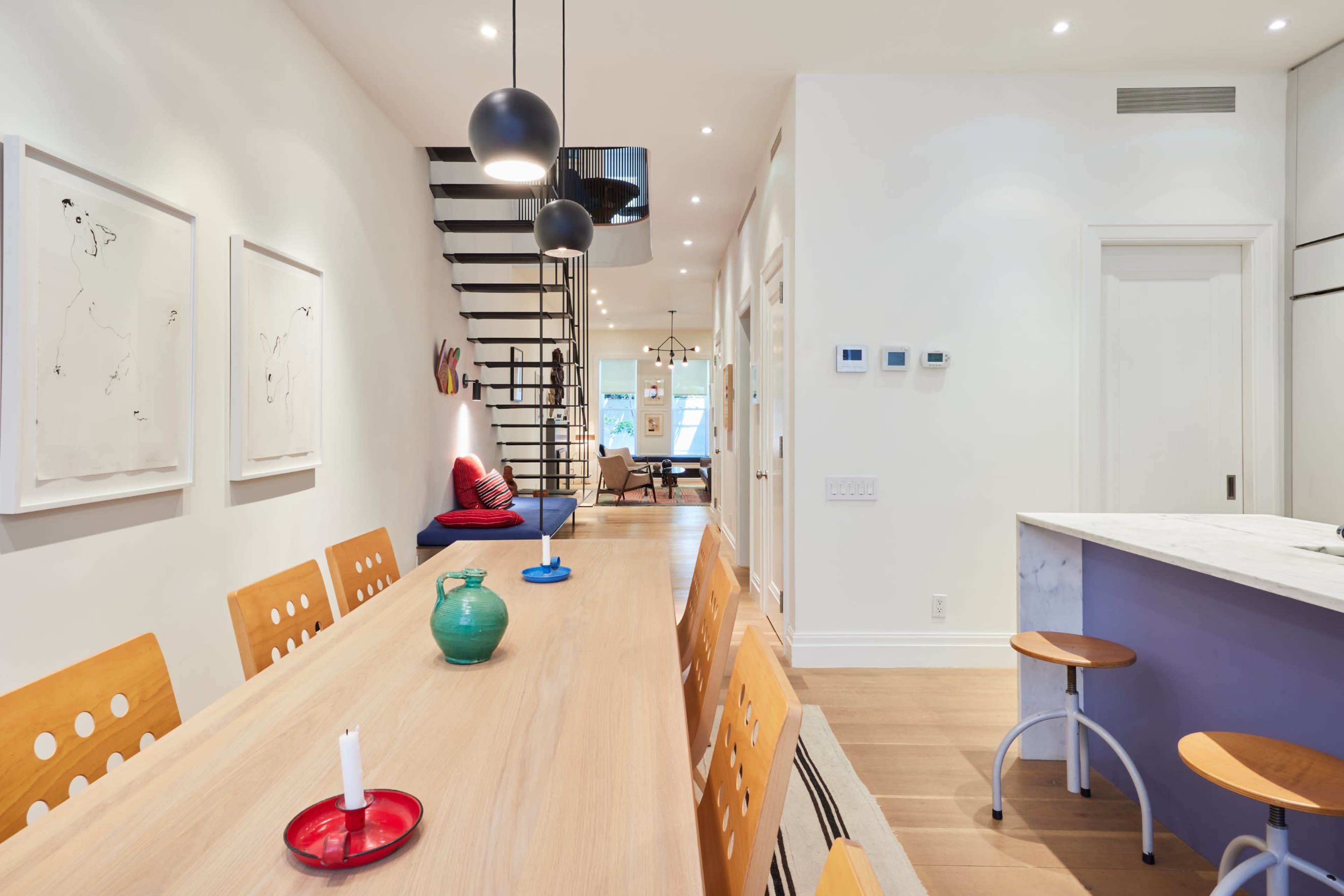 A modern dining area features a long wooden table with chairs, pendant lights overhead, and a staircase leading to an upper level, with a view of a living space in the background.