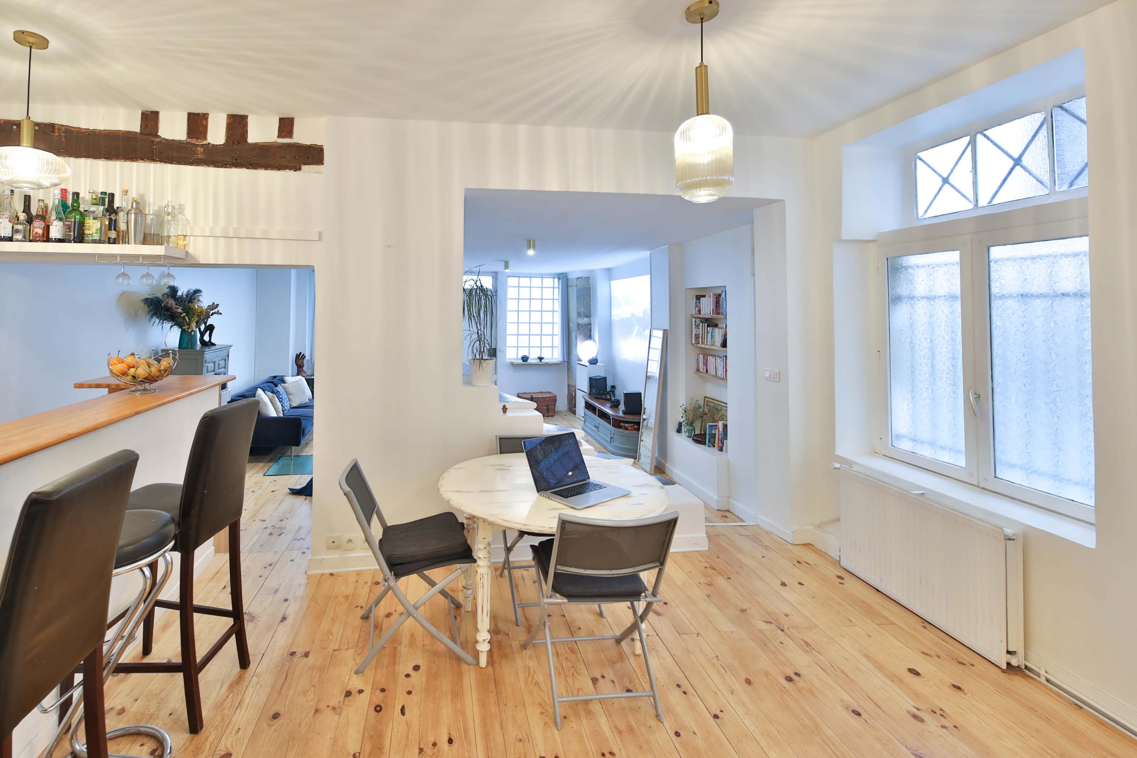 A dining area with a circular table, four chairs, and a laptop is visible next to a bar counter, leading into a well-lit living space with windows.