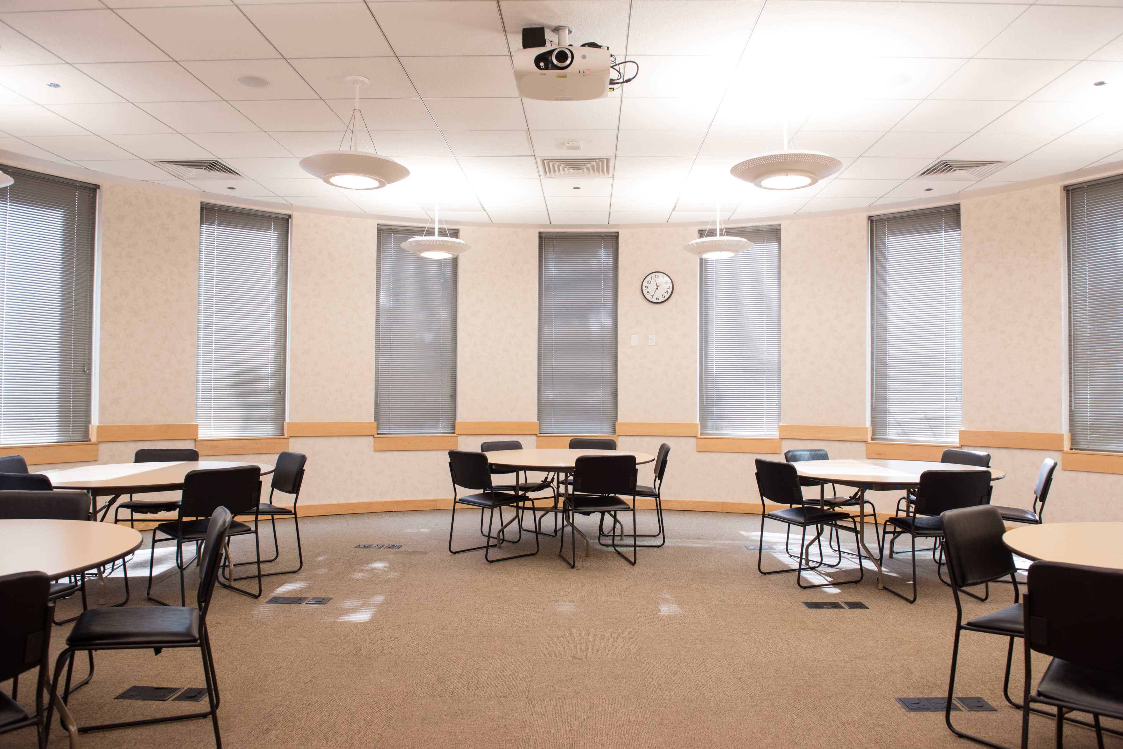 A brightly lit meeting room features several round tables surrounded by black chairs, with large windows covered by blinds.