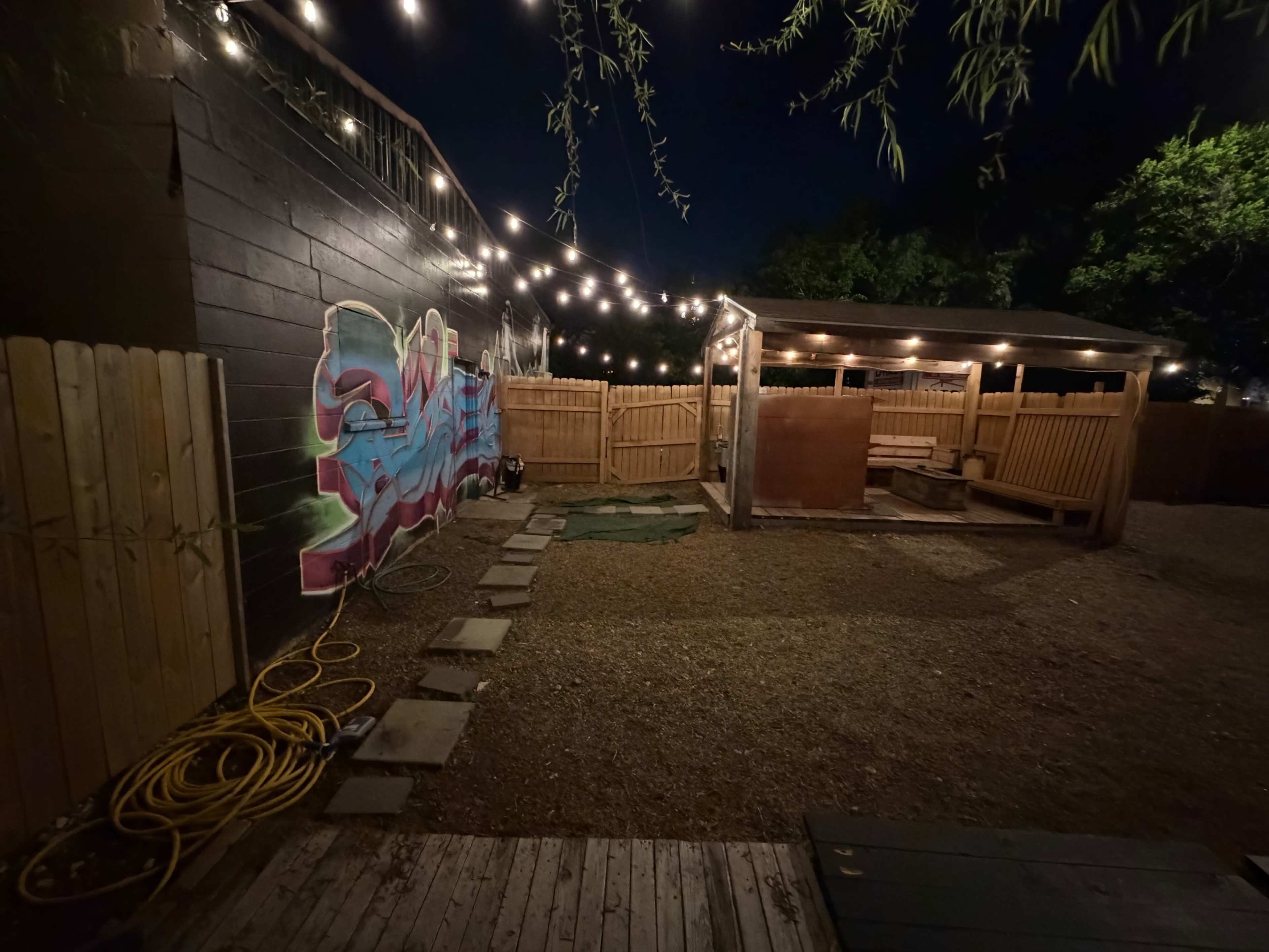 The image shows a dimly lit outdoor backyard featuring a mural on a fence, a wooden structure with seating, and string lights overhead.