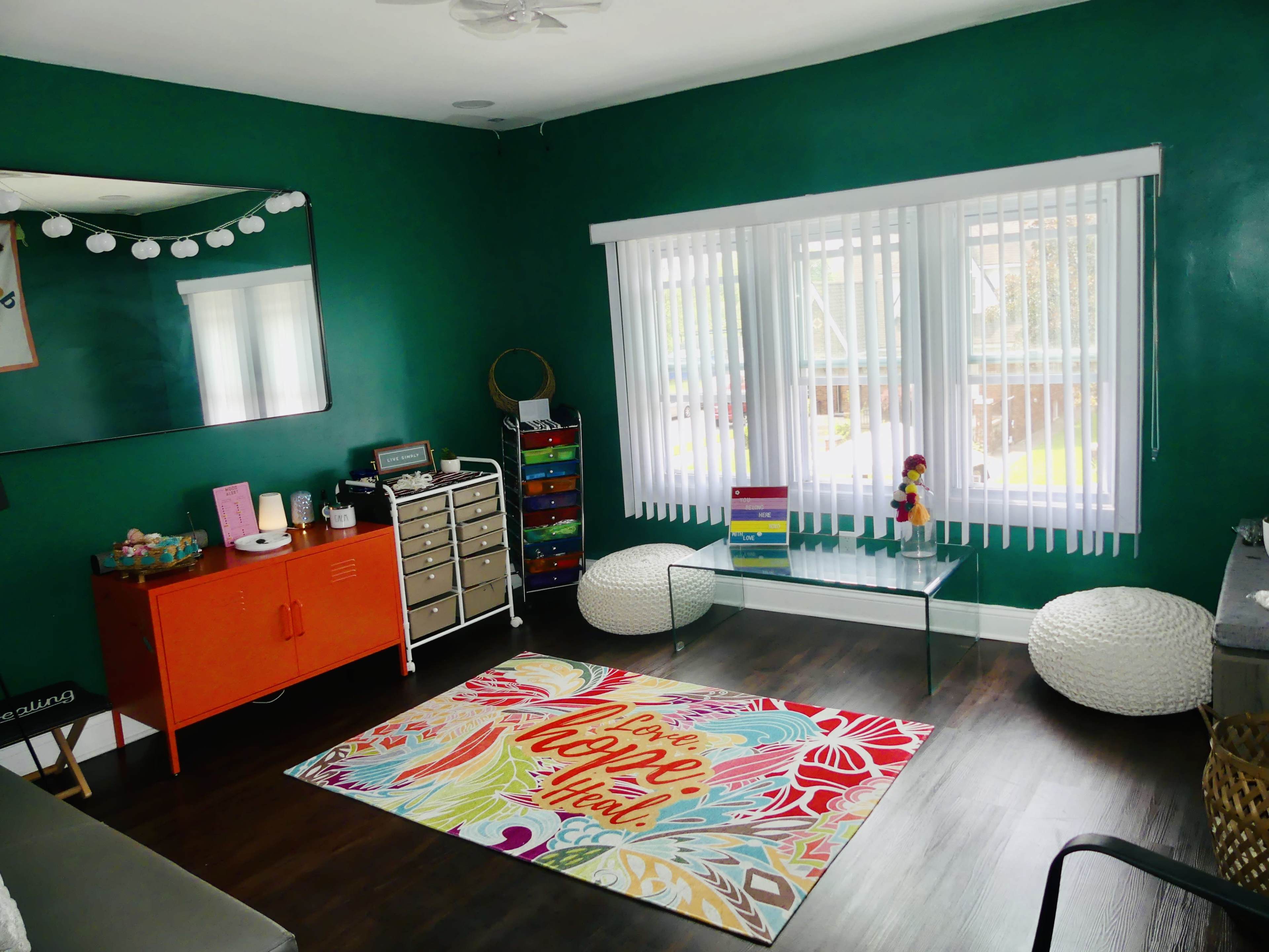 The room features green walls, a large window with white blinds, a colorful rug, and various furniture pieces including a glass coffee table and an orange cabinet.