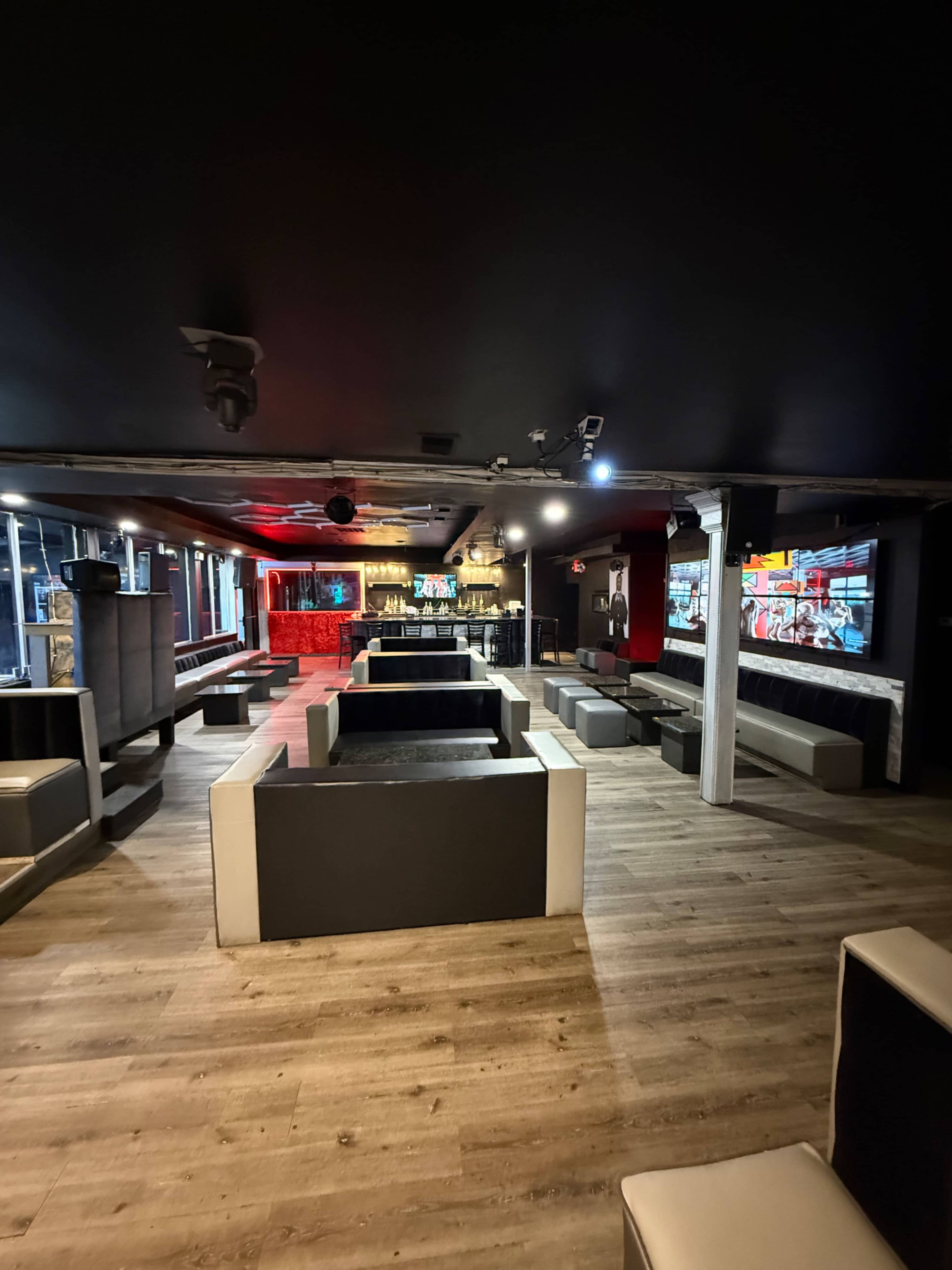 The image shows a spacious, modern lounge area equipped with low seating and a bar, featuring a dark ceiling and light-colored wooden flooring.
