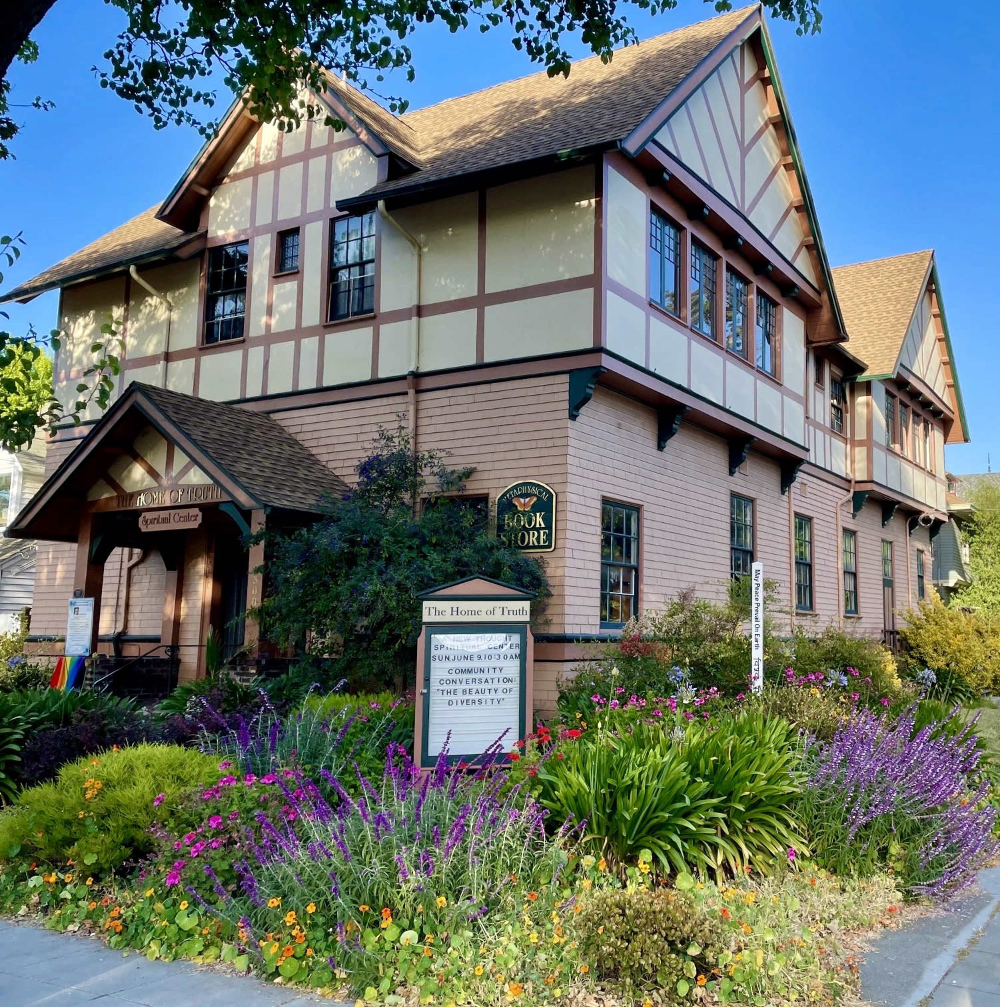 A large, two-story building with a steeply pitched roof is surrounded by colorful flowers and greenery.