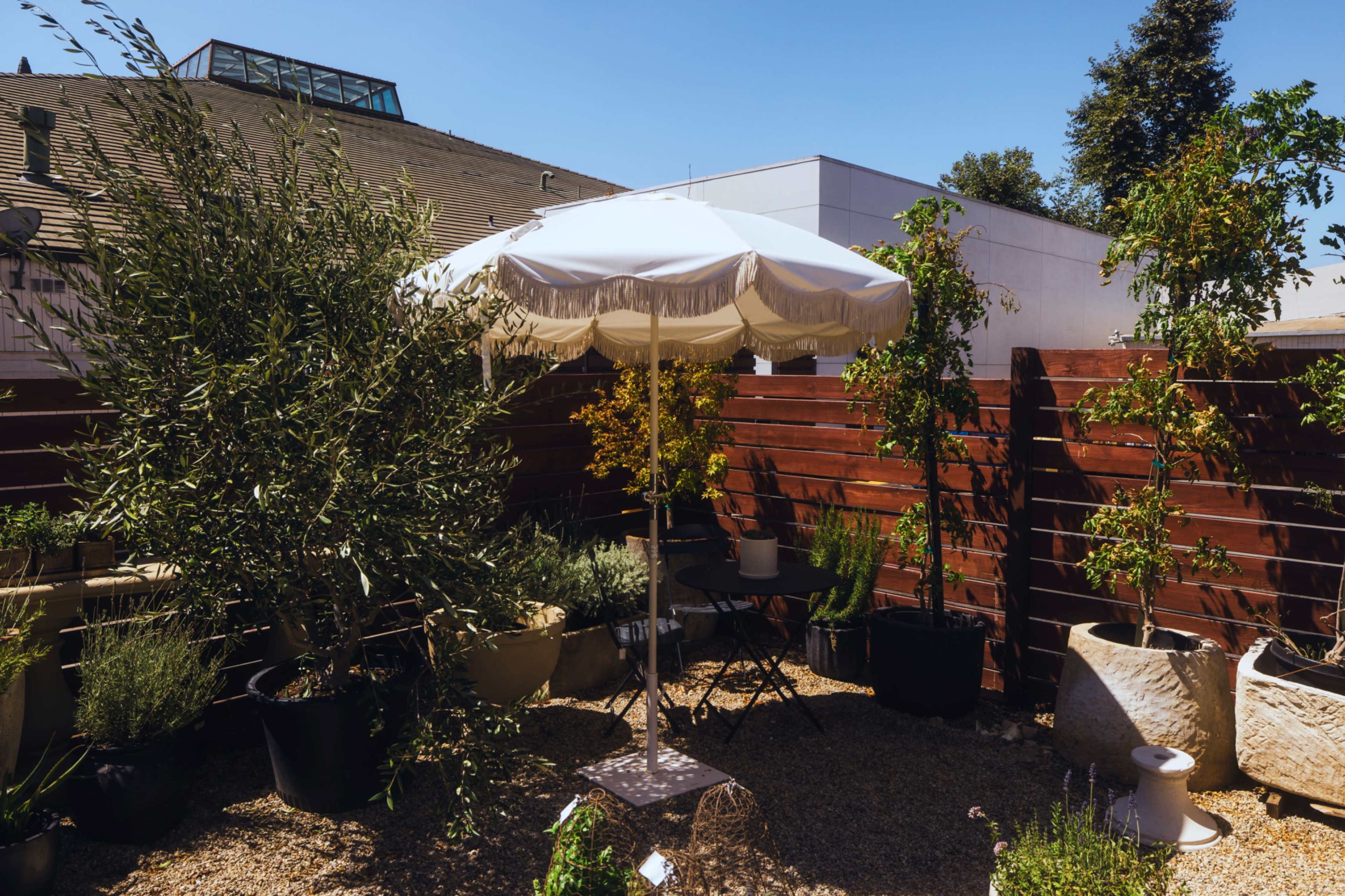 A white umbrella stands over a small table surrounded by potted plants in a gravel courtyard.