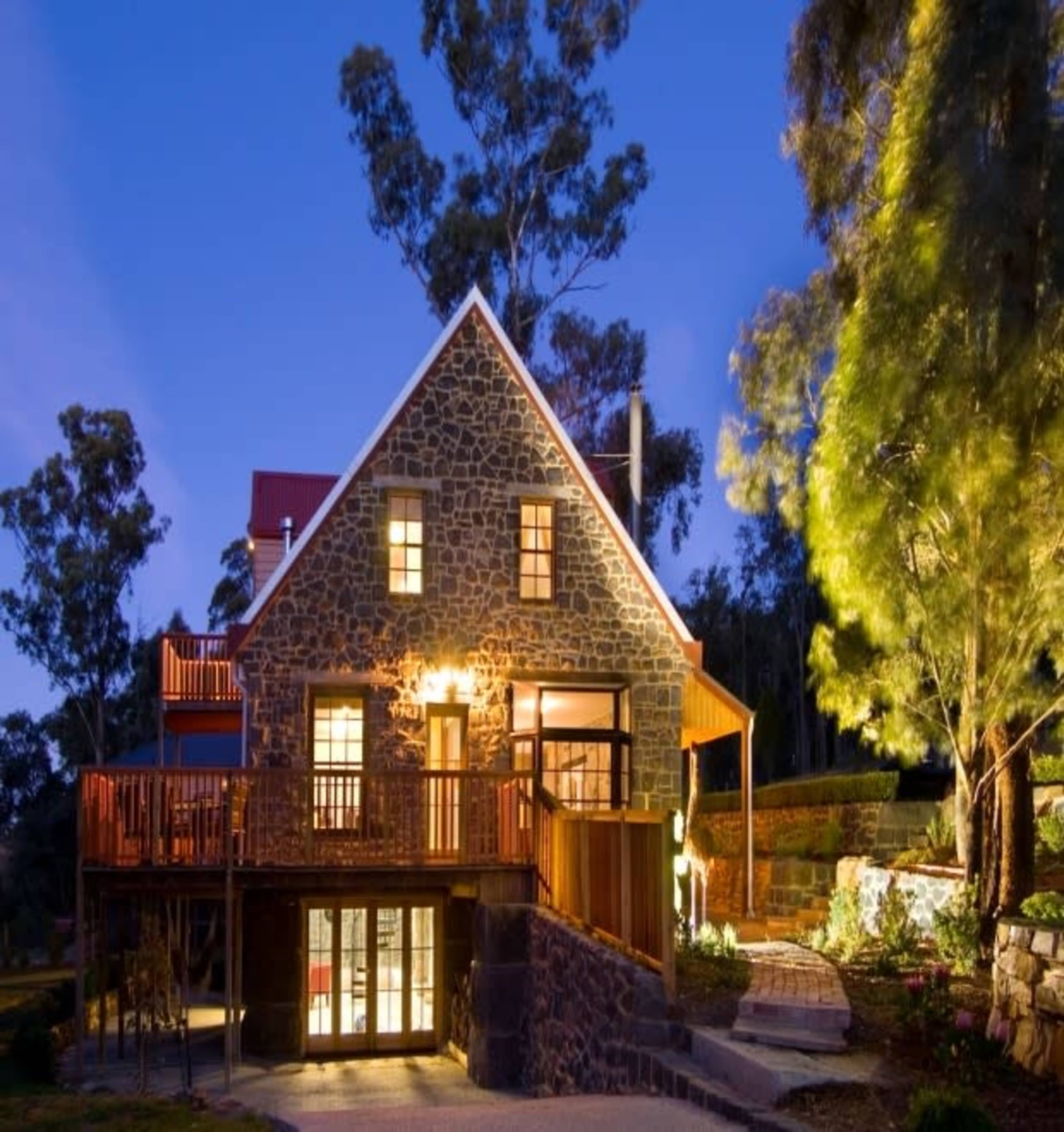 A stone house with a triangular roof and large windows is illuminated at dusk, surrounded by trees and outdoor landscaping.
