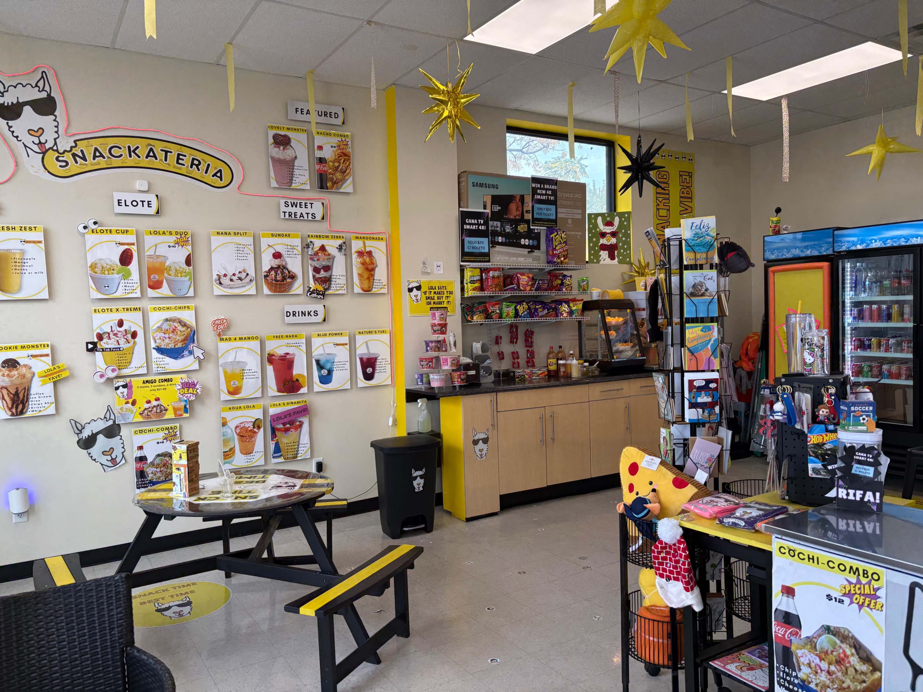 The image depicts a vibrant snack shop interior with a brightly colored menu on the wall, displaying various food and drink options, alongside a counter filled with snacks and a seating area featuring a yellow and black table.