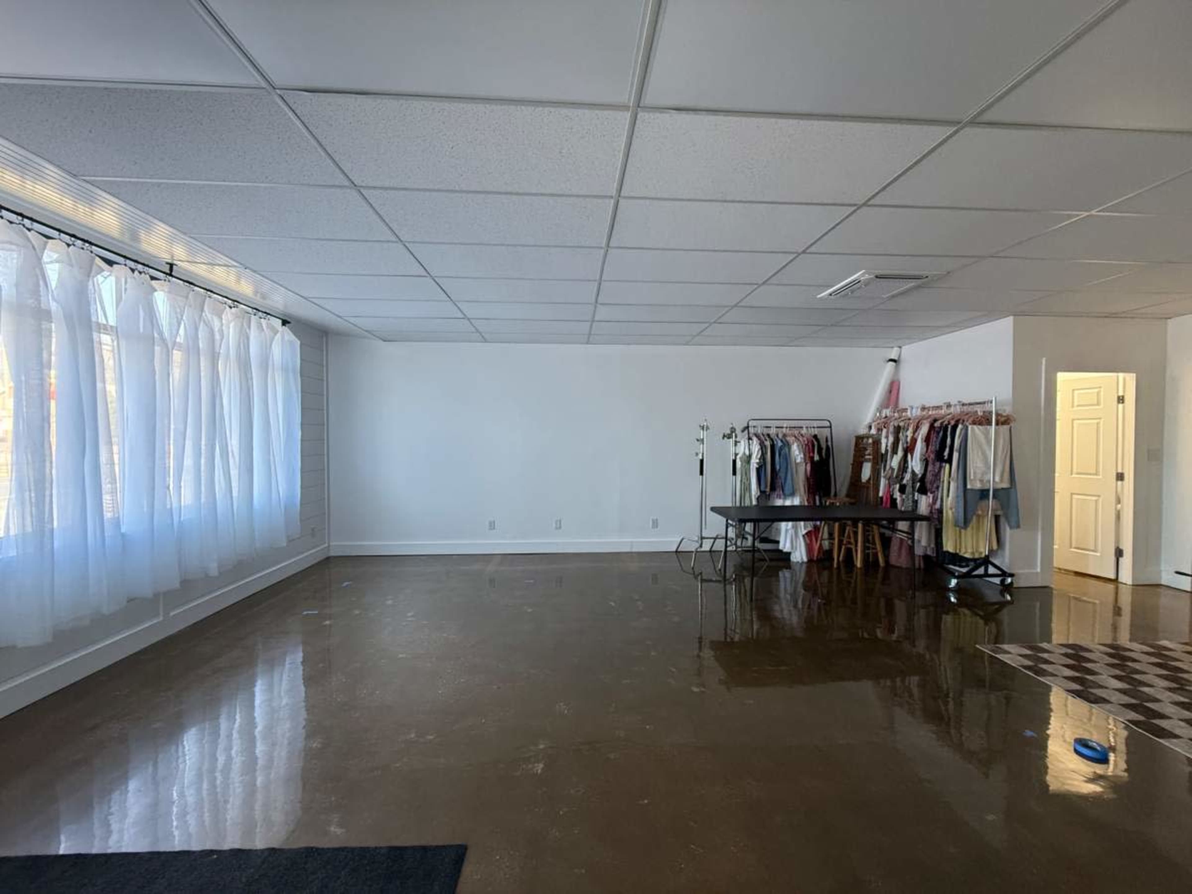 The image shows a spacious room with polished concrete flooring, large windows covered with white sheer curtains, and a row of clothing on racks along one wall, accompanied by a small table in the center.