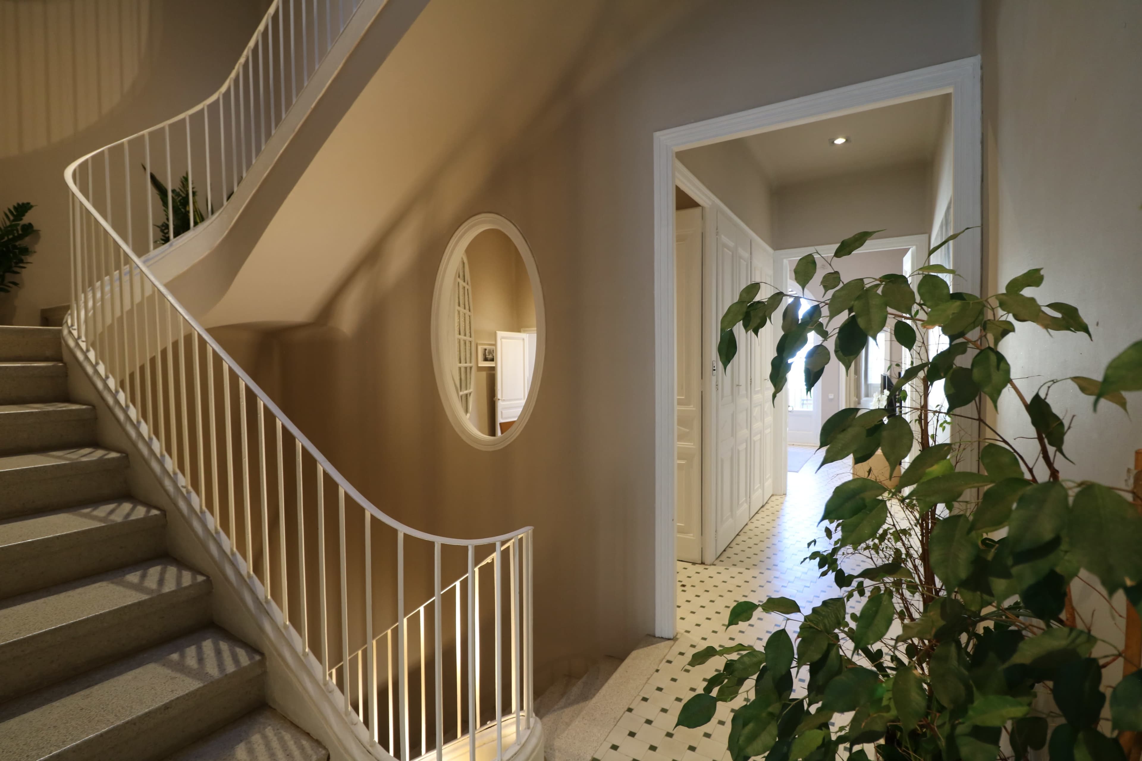 A staircase with a white railing leads to a hallway featuring an oval mirror and a floor with black and white tiles, complemented by a potted plant.