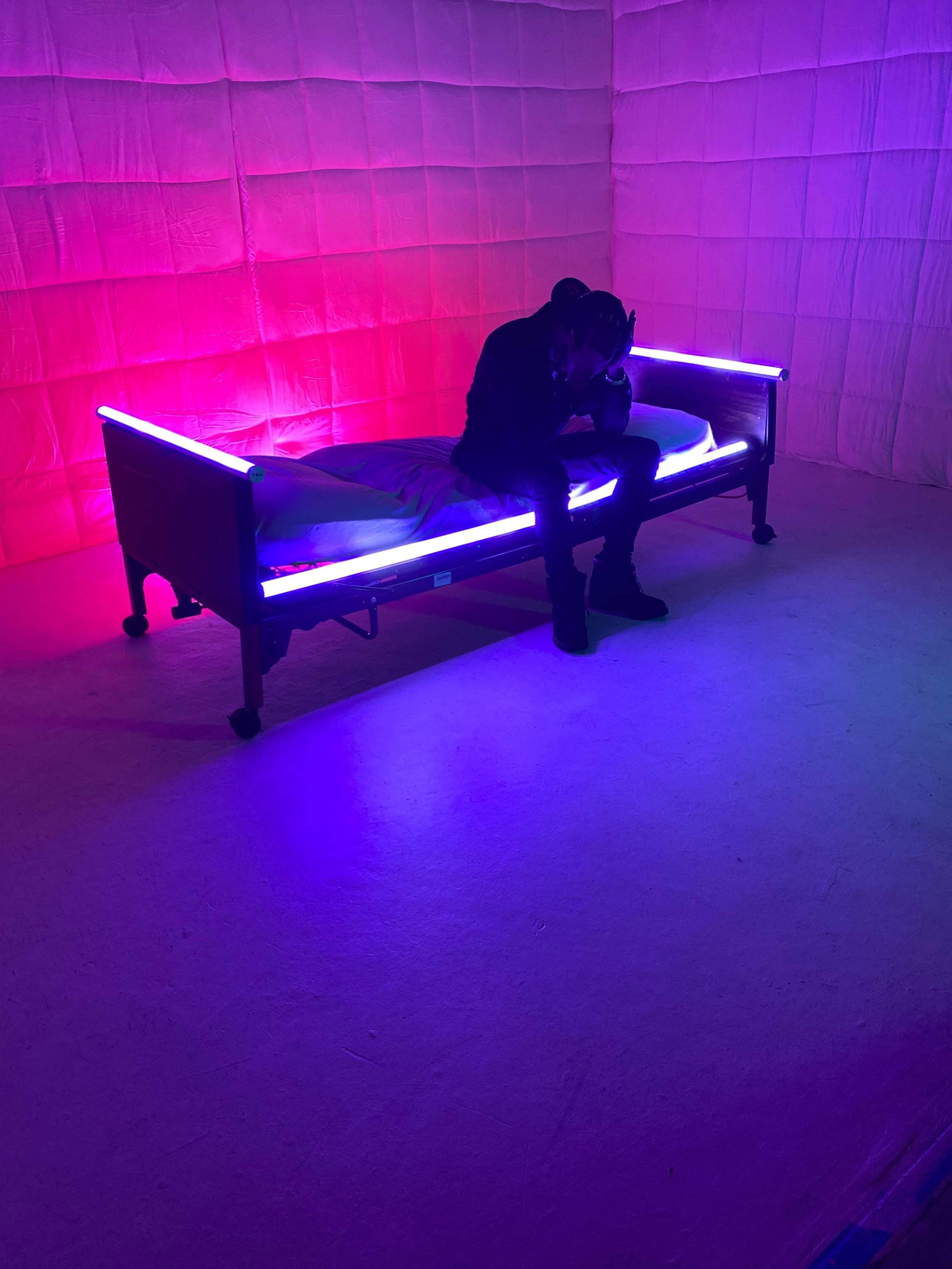 A person sits on a bed with glowing purple and pink lights in a room with textured walls.
