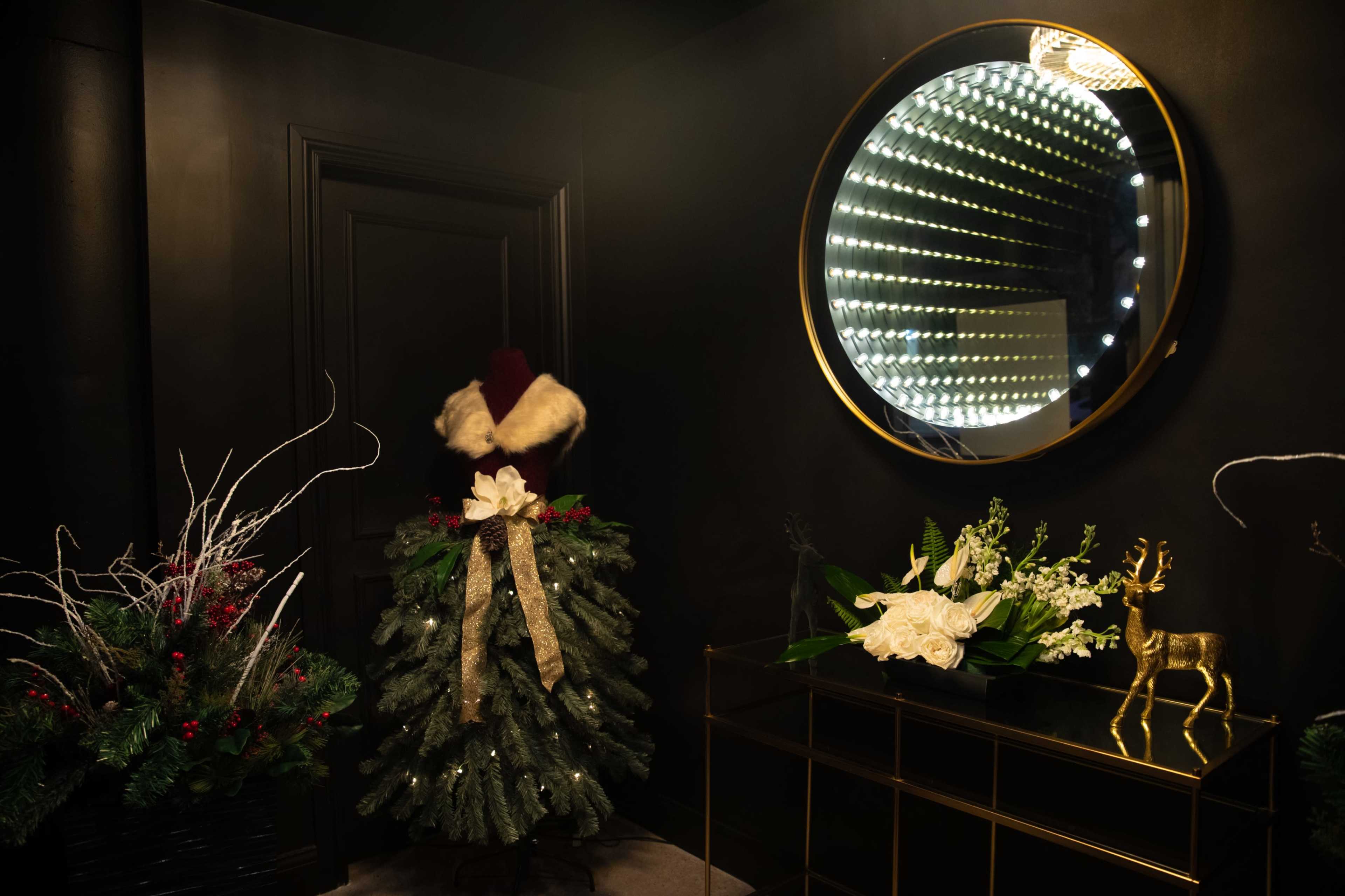 A stylish indoor scene featuring a decorated Christmas tree, a round mirror with lights, and a console table adorned with floral arrangements and a decorative golden reindeer.