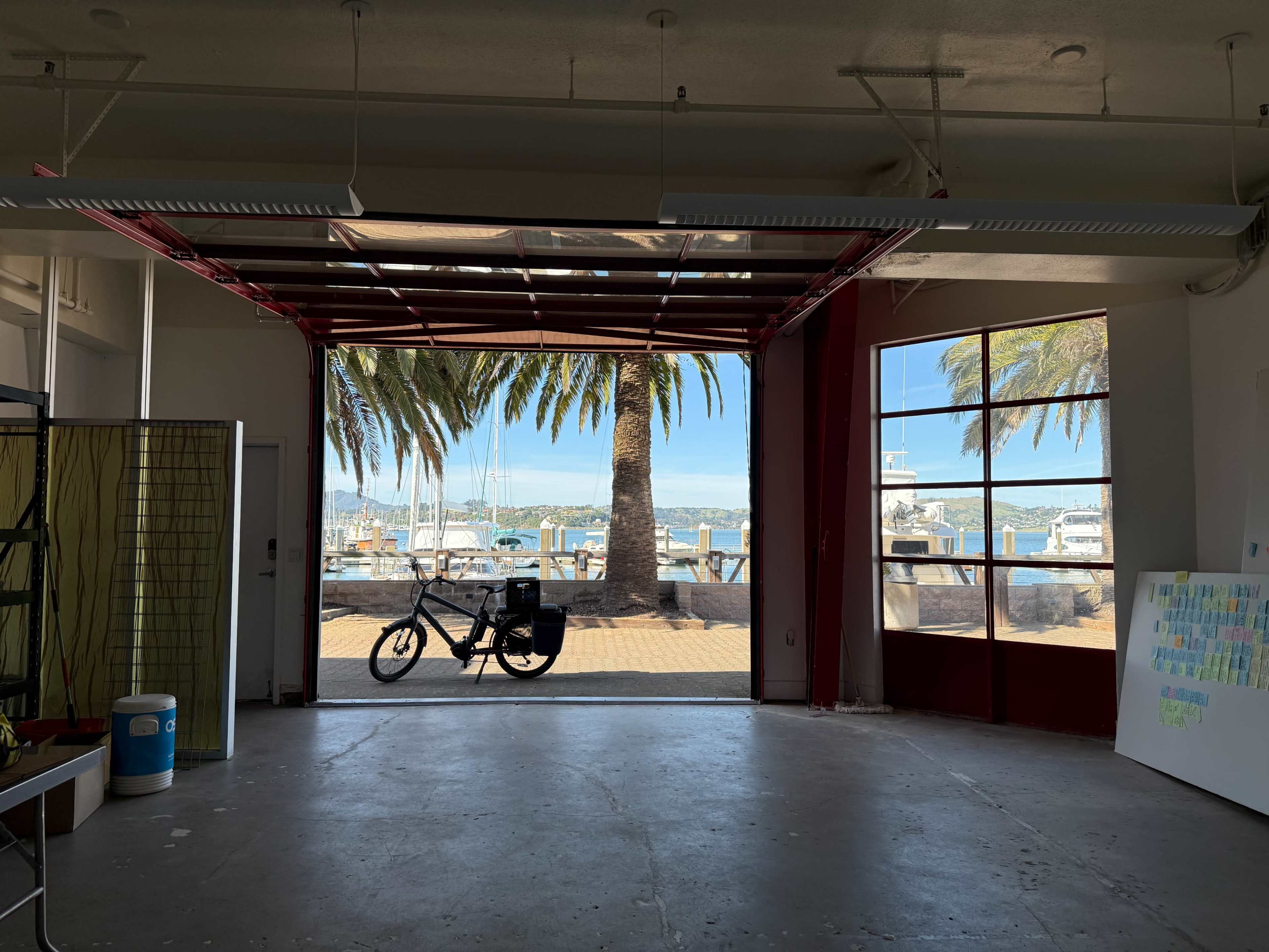 The image shows a spacious room with large openings leading to a waterfront view, framed by palm trees and boats in the background.