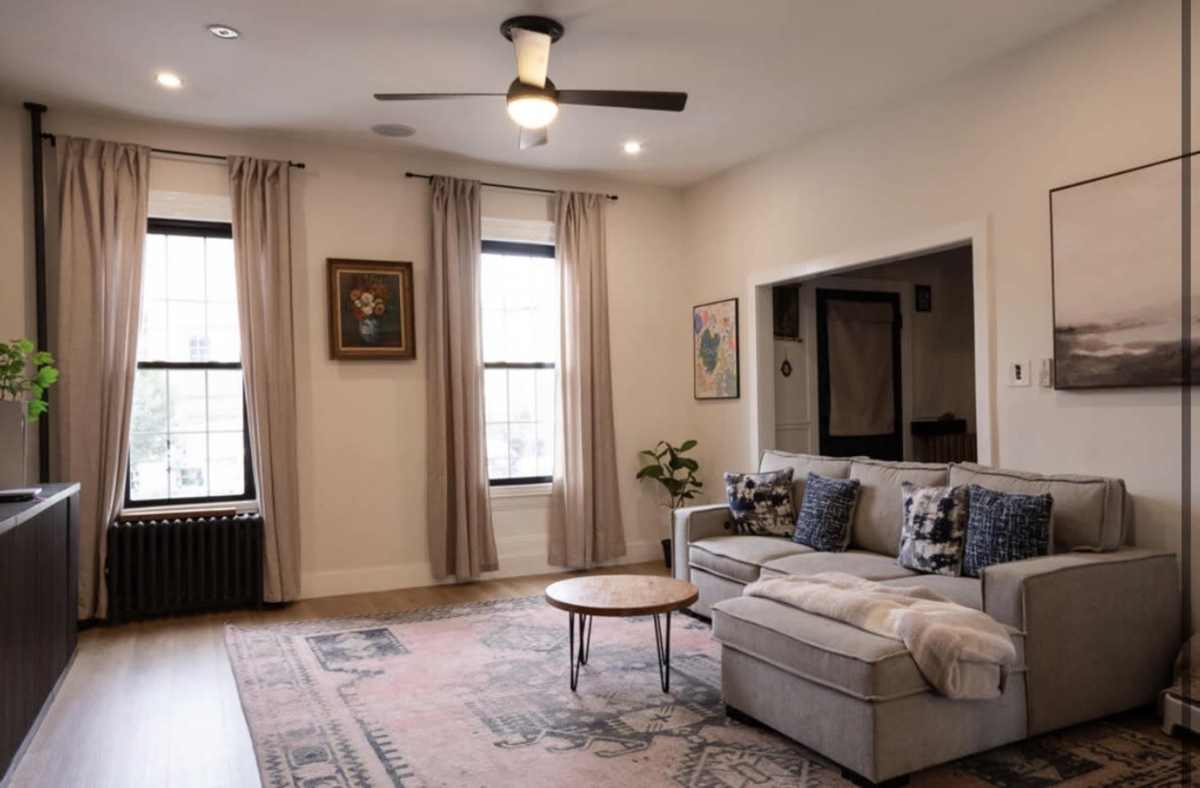The image shows a spacious living room with a light-colored sofa, a round coffee table, large windows with curtains, and wall art.