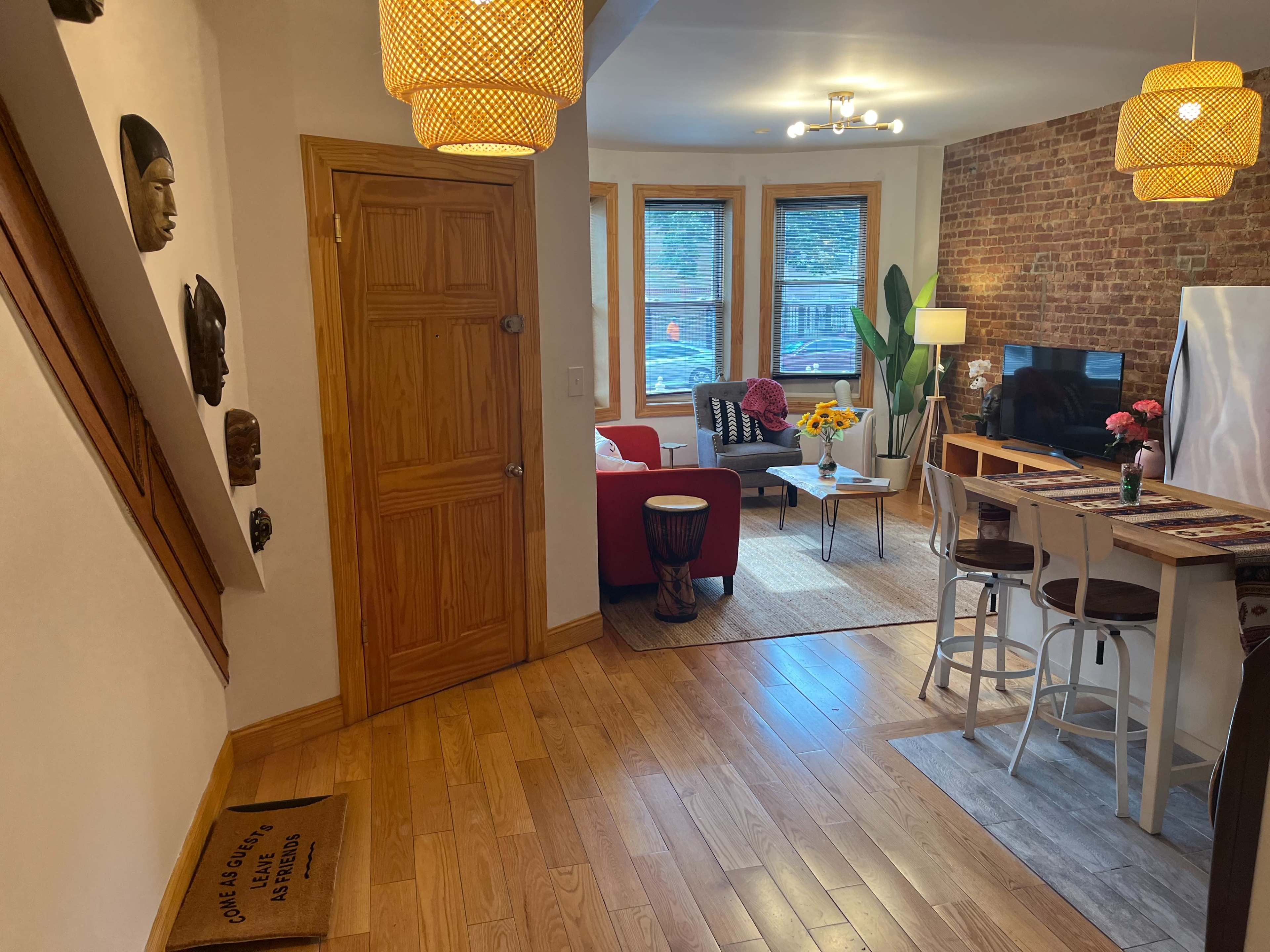 A spacious living area with a wooden floor, featuring a kitchen bar, a red chair, a television mounted on a brick wall, and decorative masks on the adjacent wall.