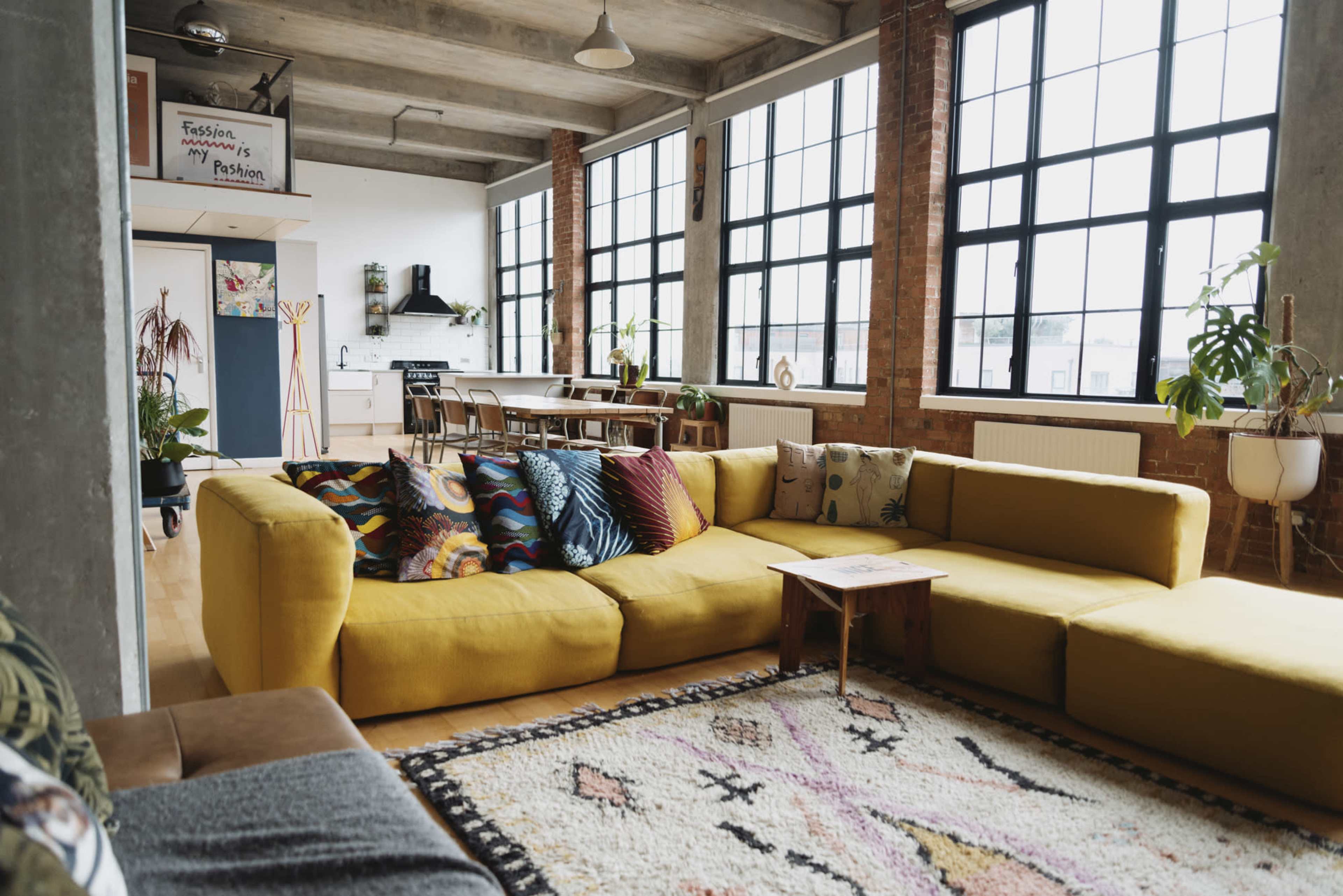 A spacious, open-concept living area with a large yellow sectional sofa, a wooden dining table, and large windows allowing natural light to fill the room.