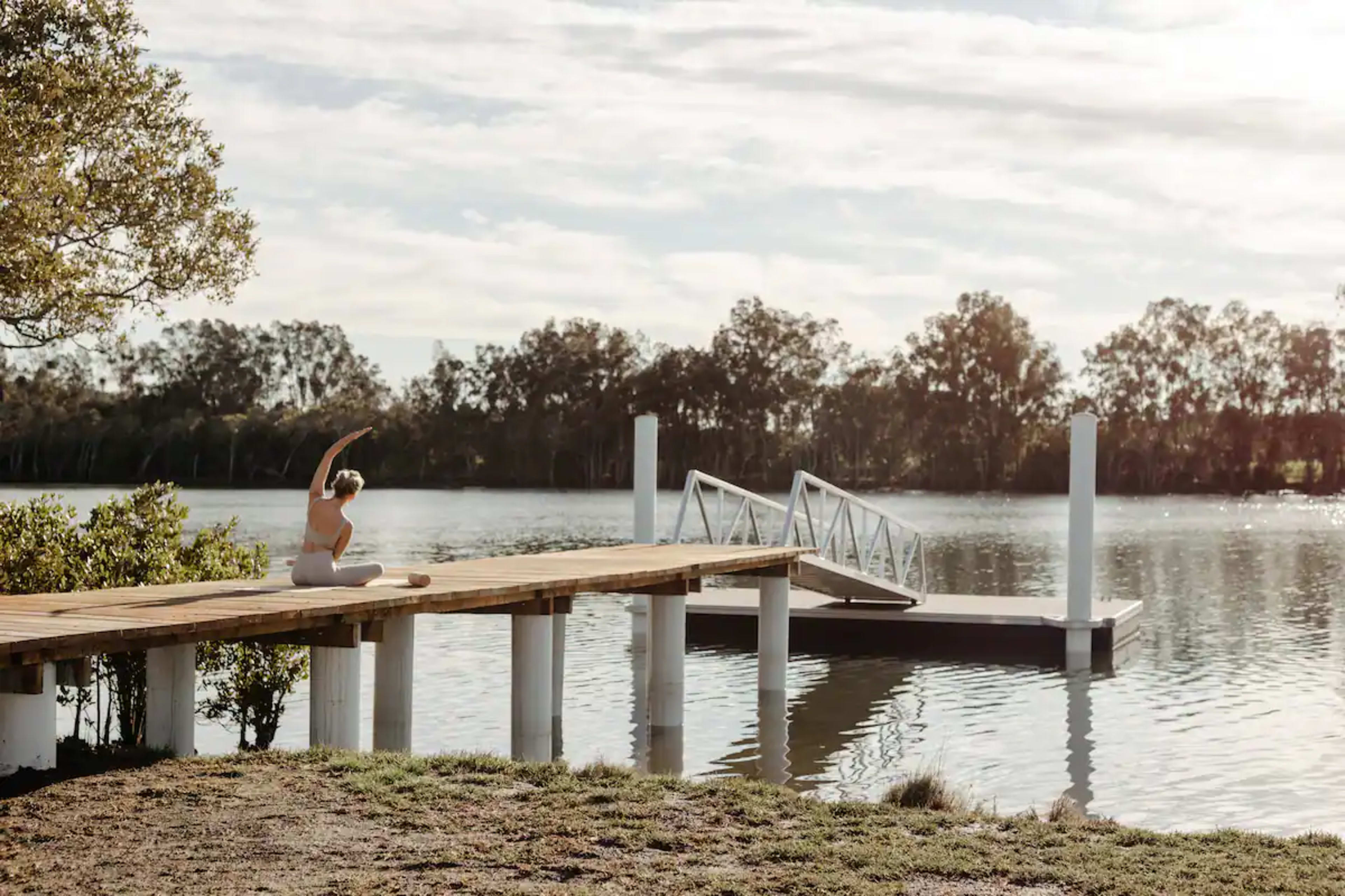 A person practices yoga on a wooden dock overlooking a calm lake surrounded by trees.