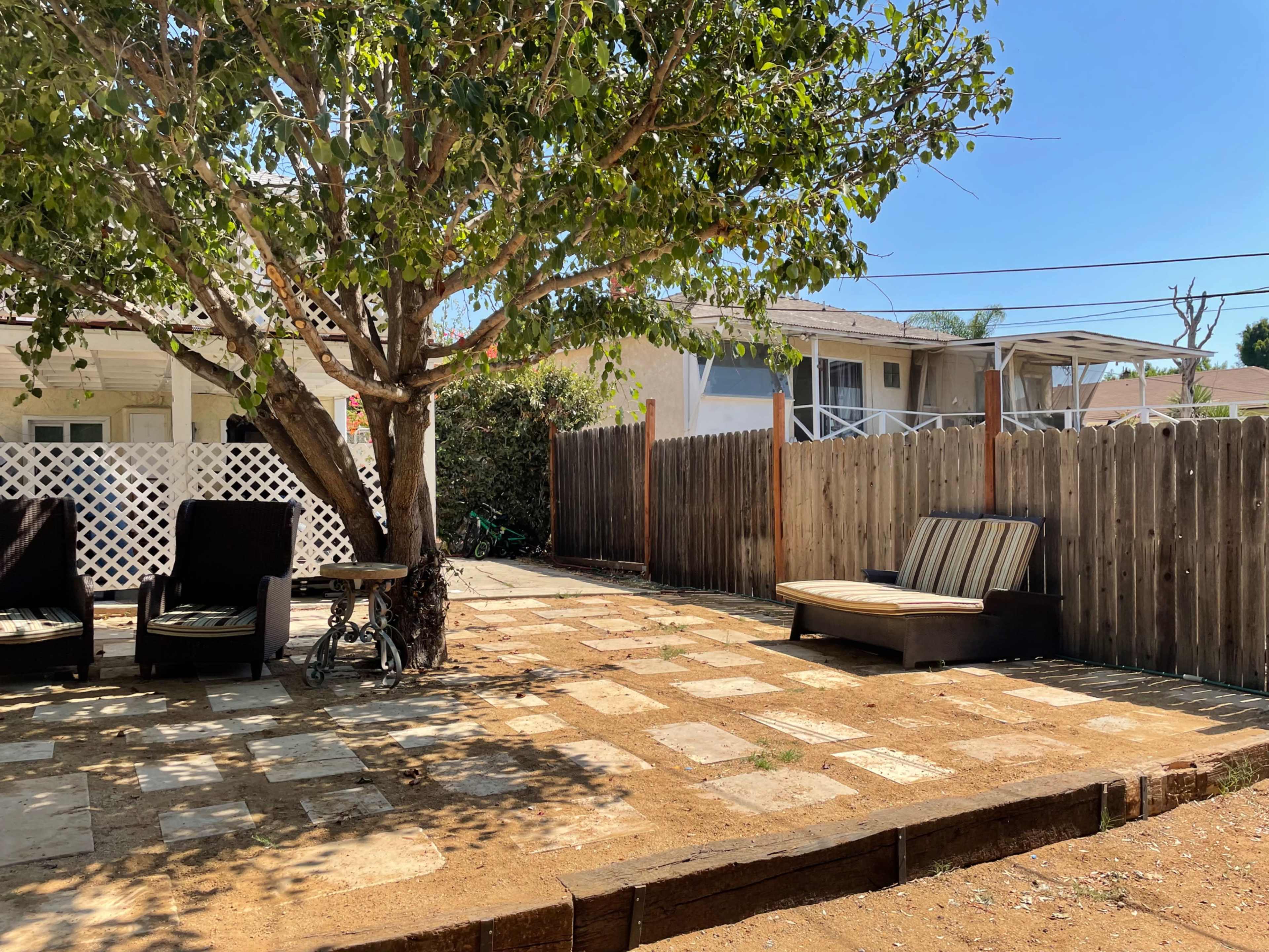 The image shows a backyard with a tree, two lounge chairs, and a stone patio surrounded by a wooden fence.