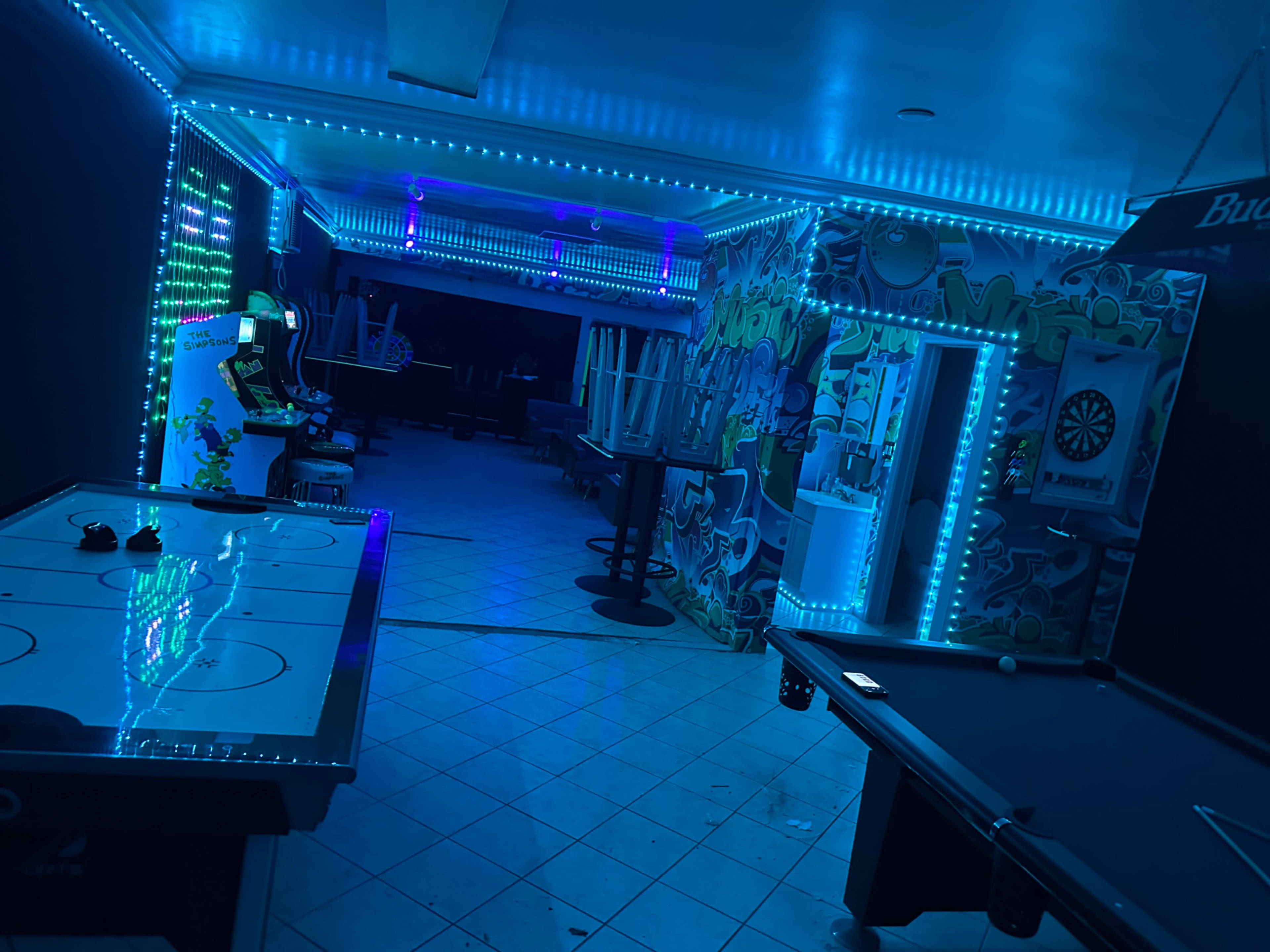The image shows a dimly lit game room featuring an air hockey table, a pool table, and neon blue lighting along the walls.
