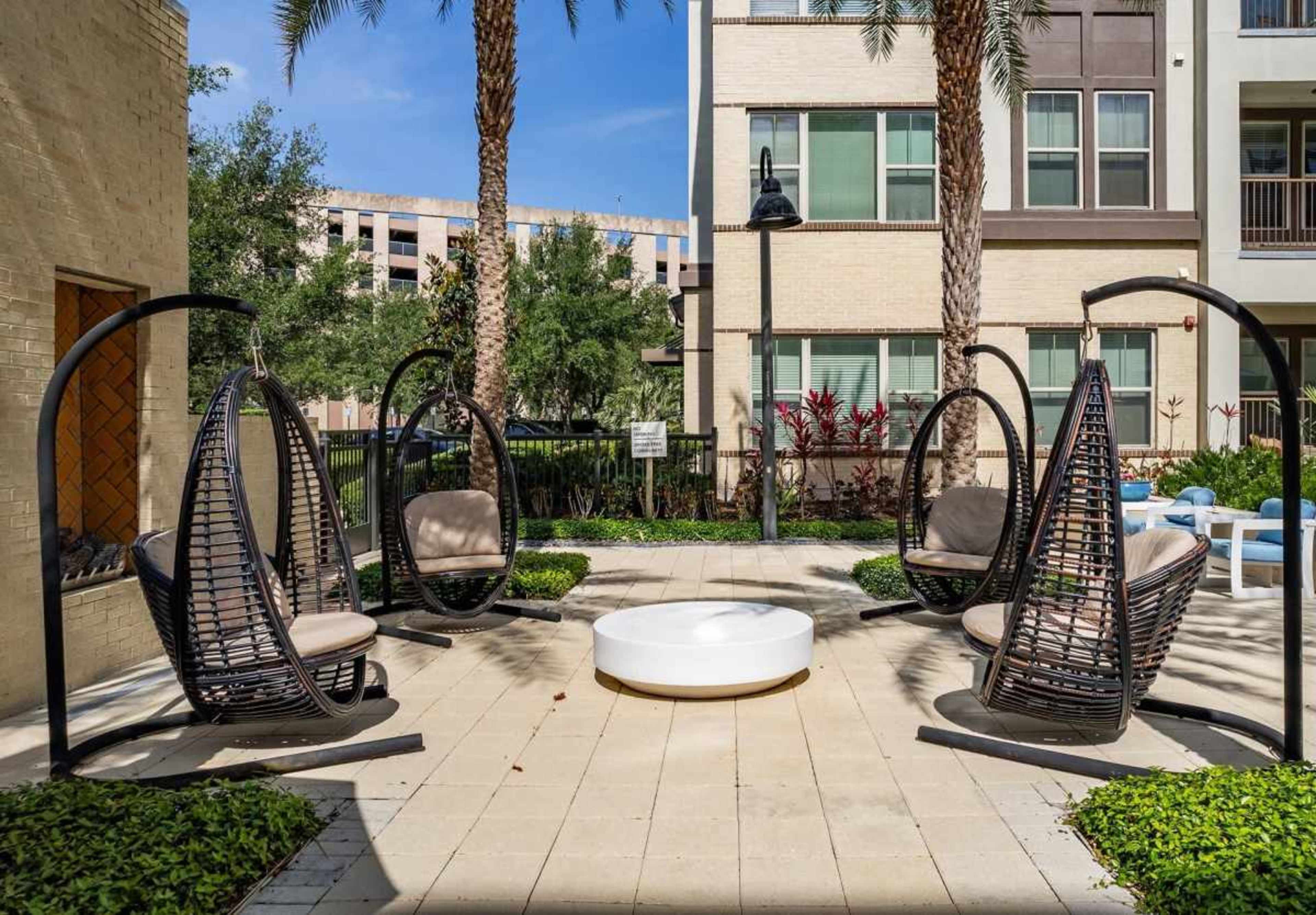 The image shows a seating area with four hanging chairs arranged around a circular fire pit in a courtyard surrounded by greenery and apartment buildings.