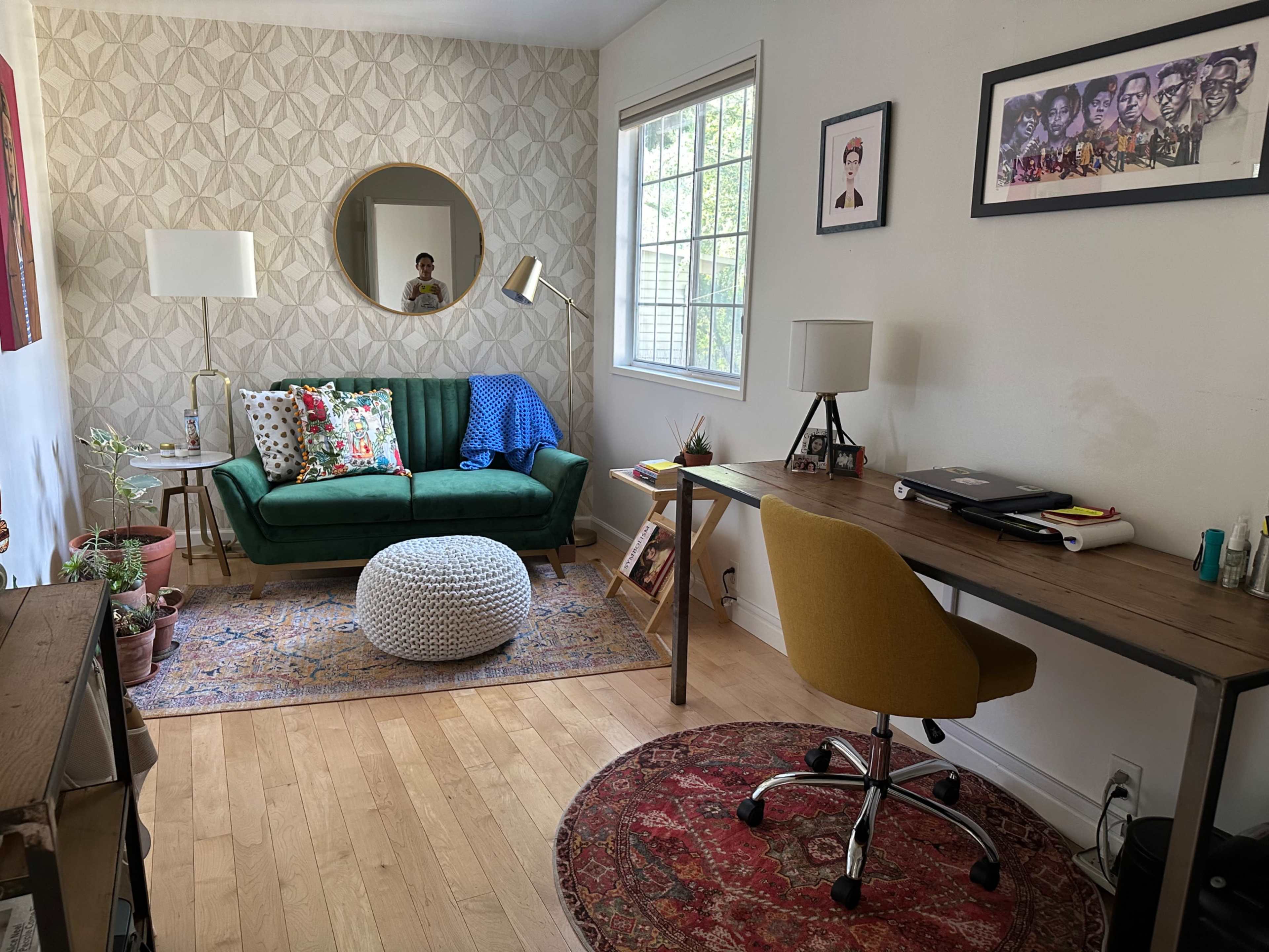 The image shows a cozy home office with a green sofa, a round mirror, a wooden desk with a chair, and various decorative elements including plants and artwork on the walls.