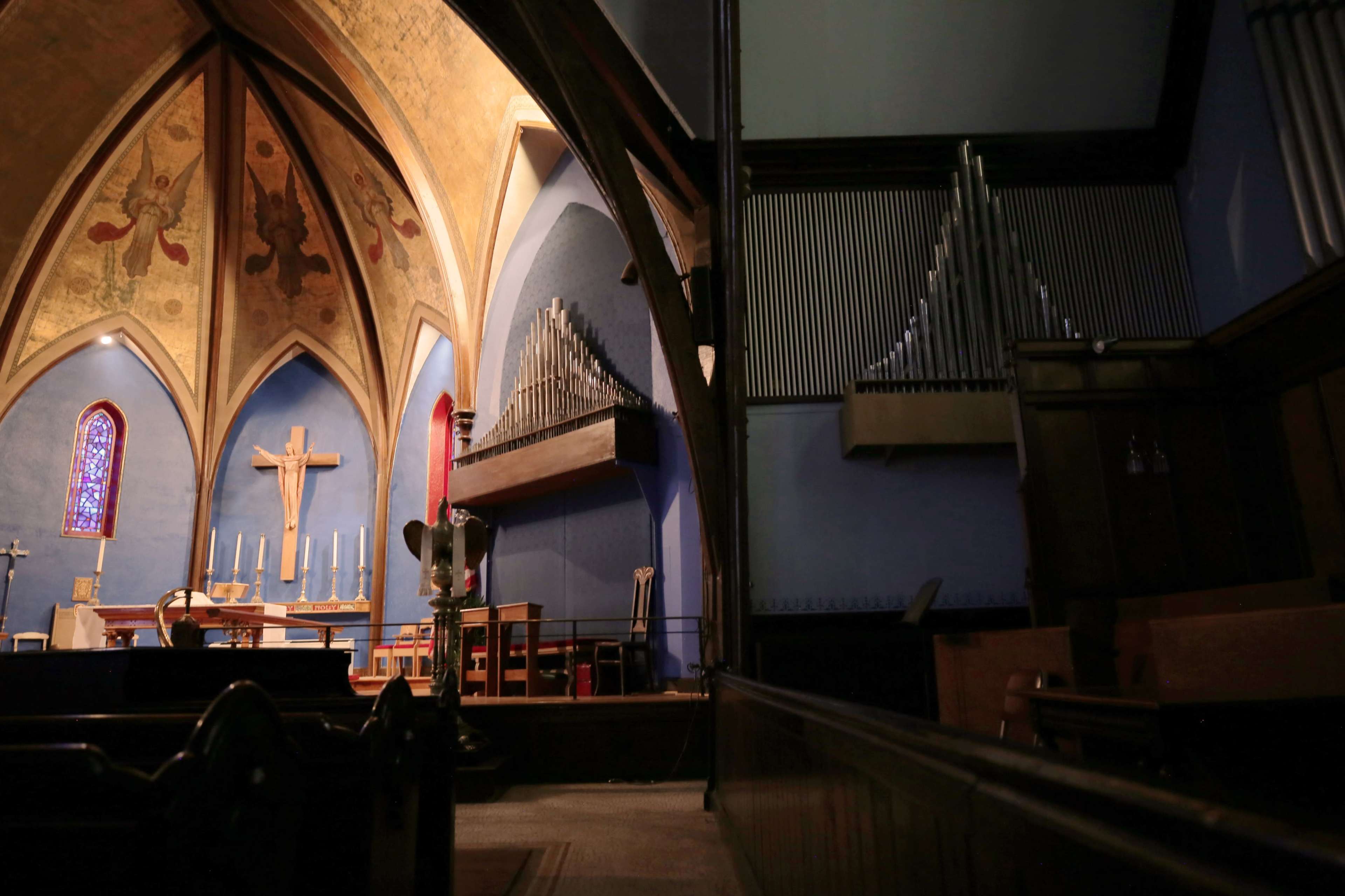 The interior of a church features a raised altar with a wooden cross, flanked by multiple pipe organs and stained glass windows.