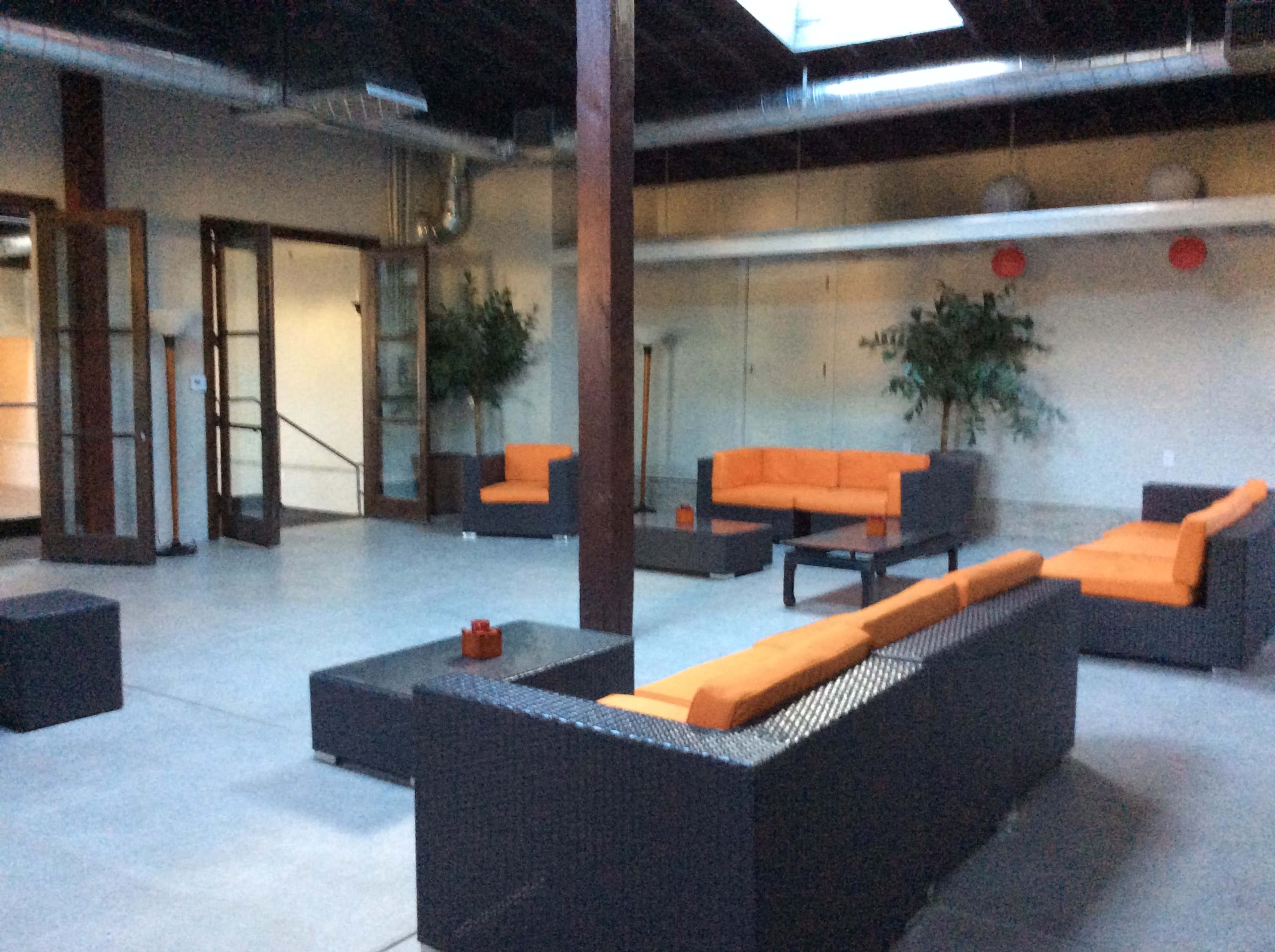 The image shows a spacious room with modern furniture, featuring orange-cushioned seating arrangements and a central coffee table in a well-lit environment.