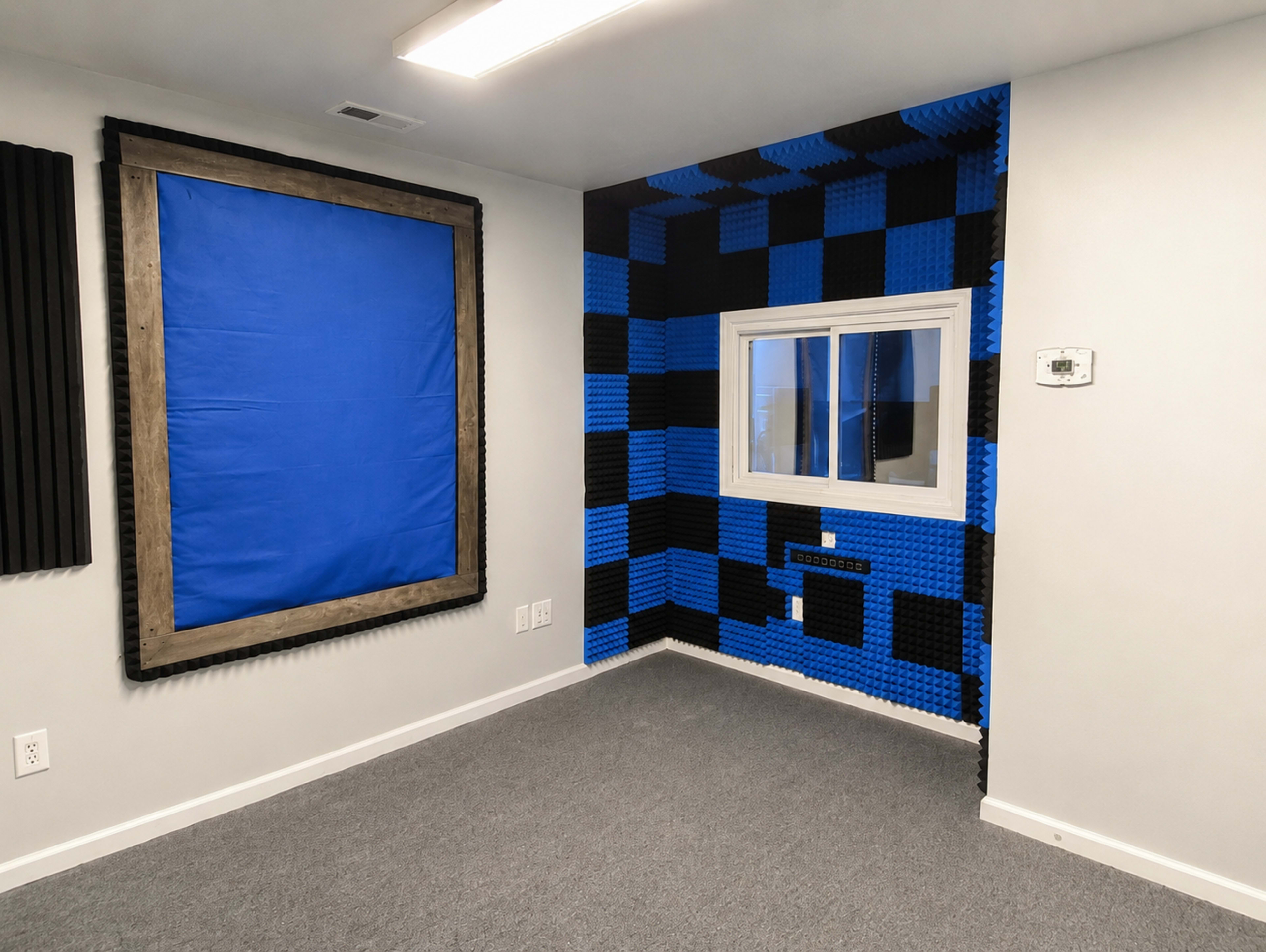 The image shows a room with blue and black soundproofing panels on the walls, a framed blue cover over a window, and a gray carpeted floor.