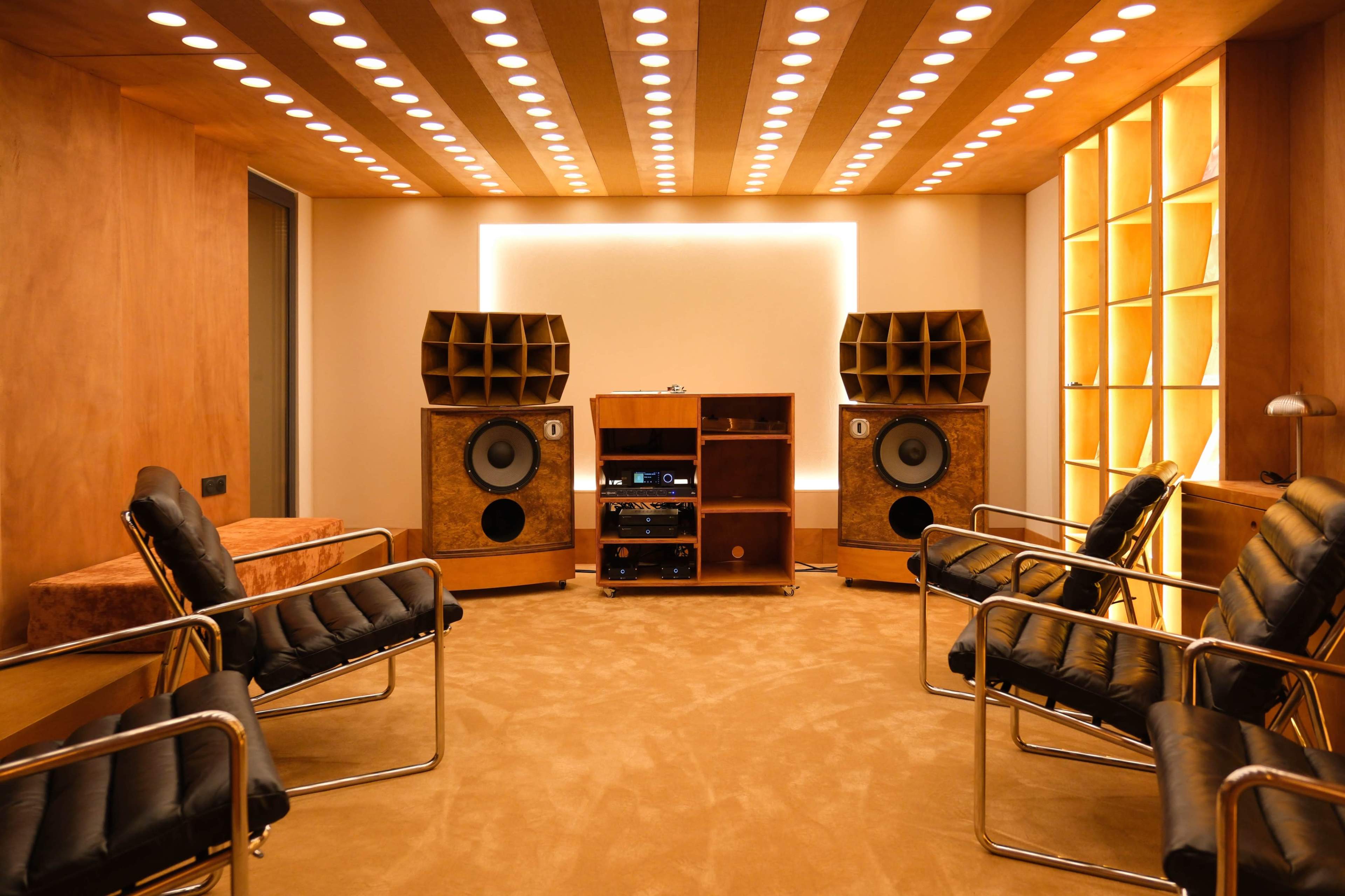 The image shows a modern room designed for audio listening, featuring two large speakers, a wooden audio rack, and several black lounge chairs arranged in front of them.