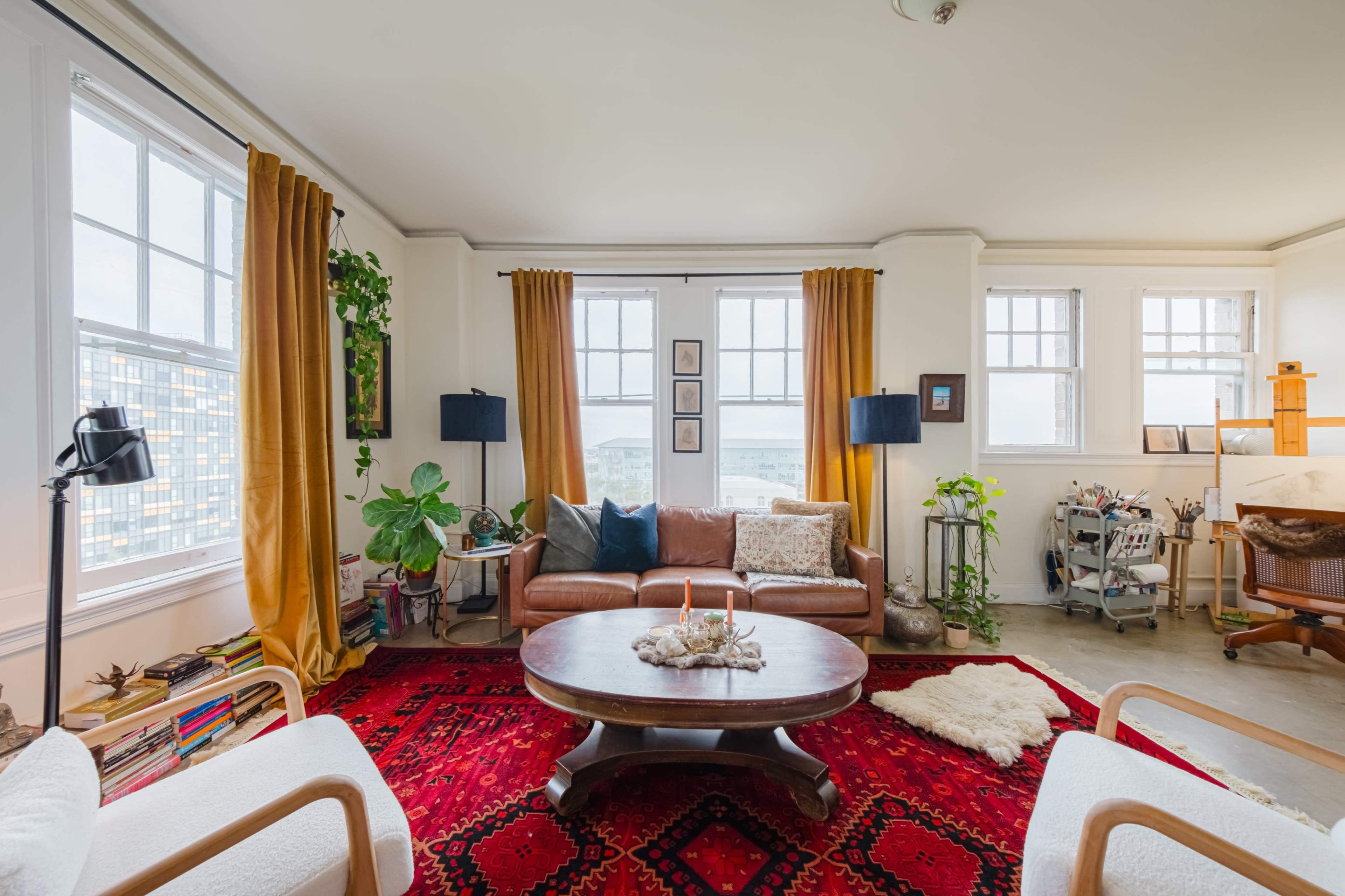 The image shows a cozy living room with a brown leather sofa, a round wooden coffee table, and large windows framed by yellow curtains.