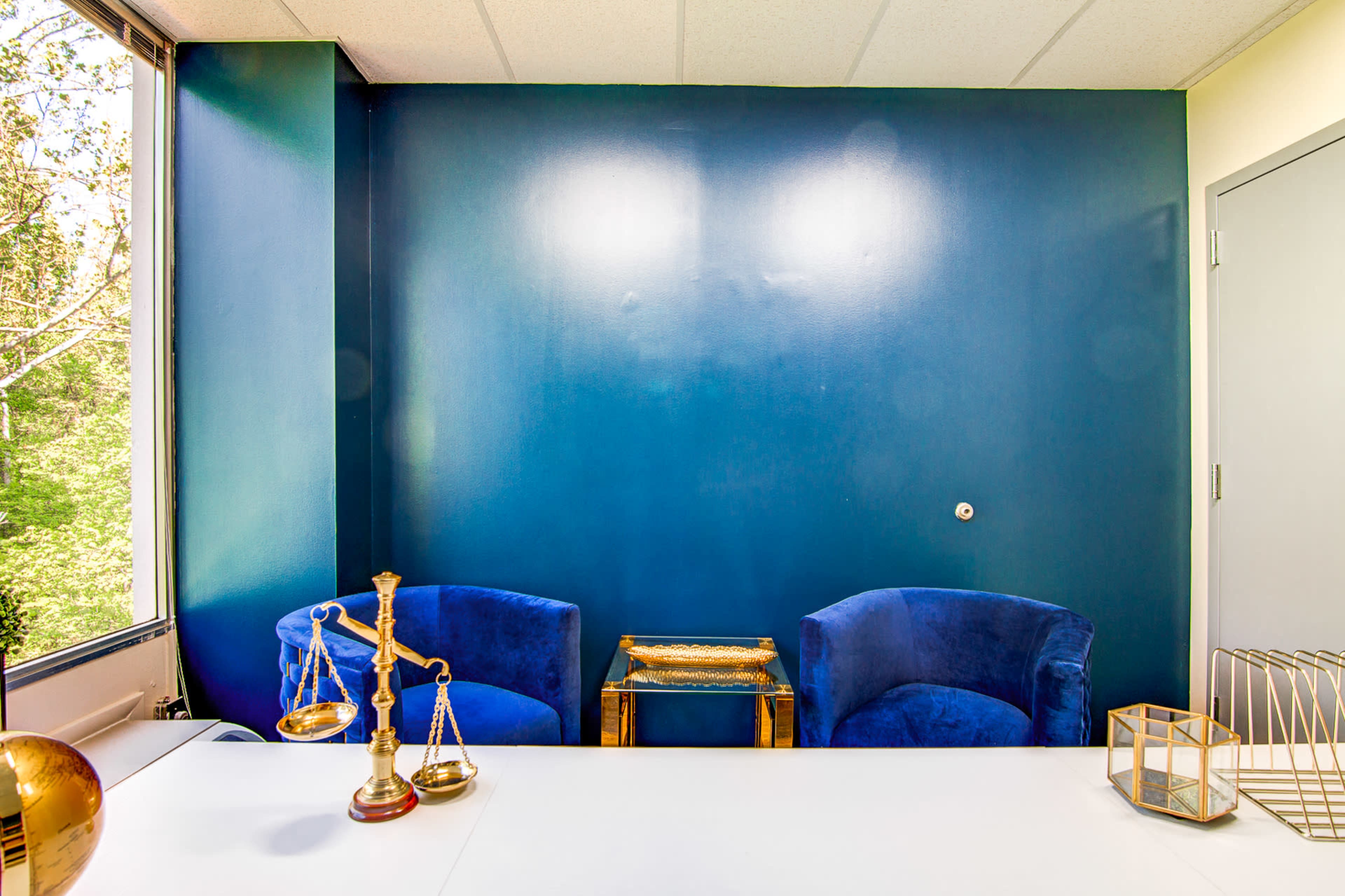 An office space with two blue chairs facing a small table against a dark blue wall, complemented by a window revealing greenery outside.