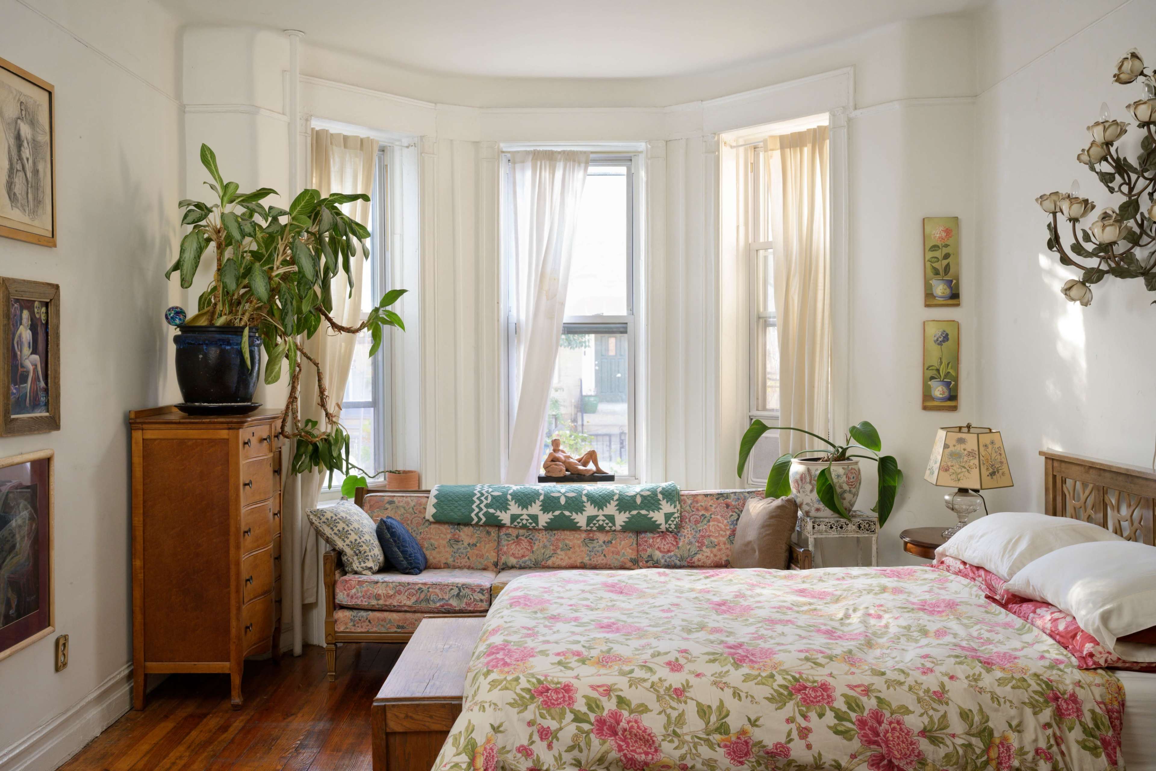 The image shows a bright, airy bedroom featuring a floral-patterned bedspread, a wooden dresser, a small couch with decorative pillows, and natural light streaming through large windows.