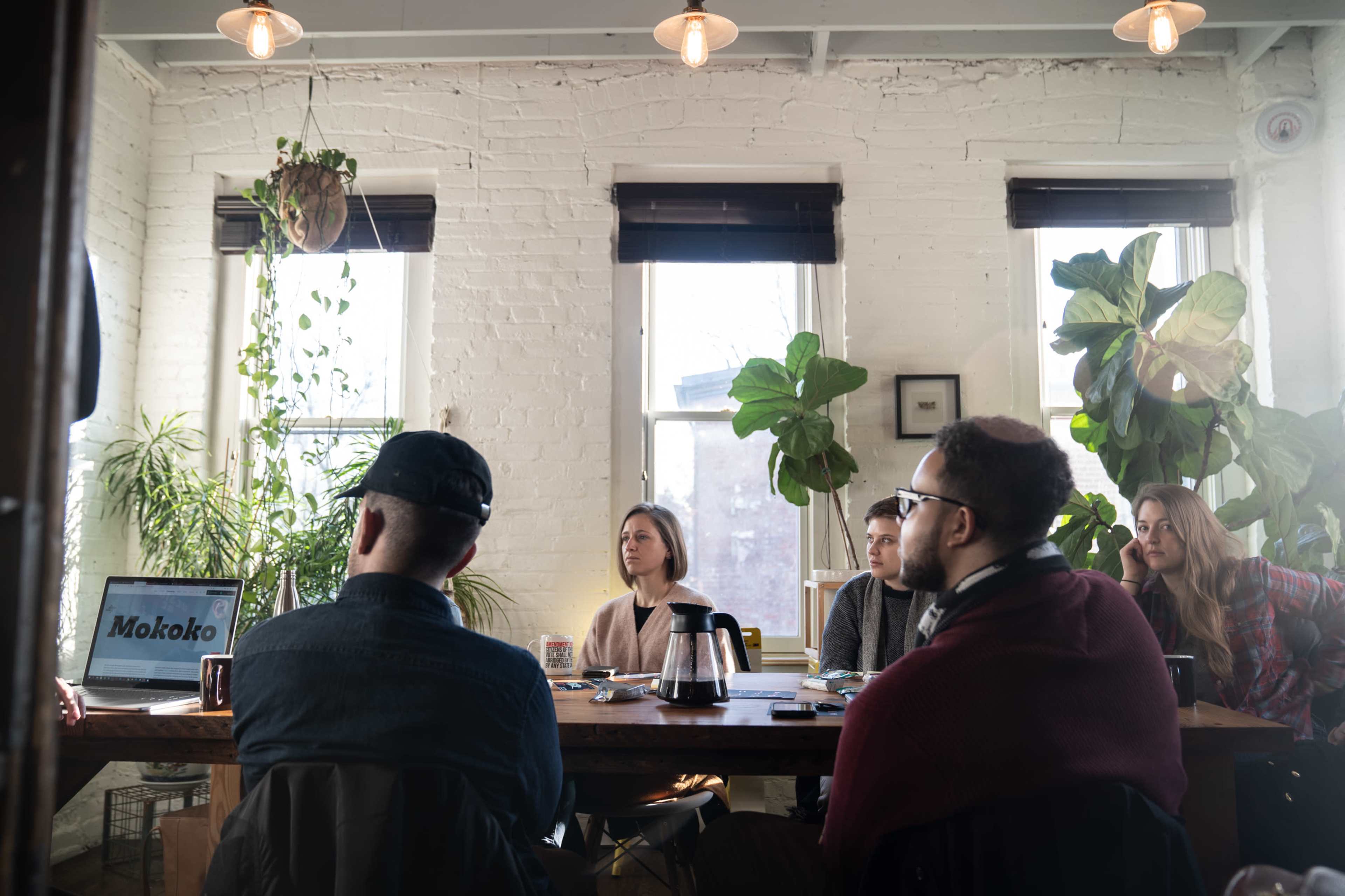 A group of six people sits around a table in a bright room filled with plants, engaged in discussion while a laptop displays the word "Mokoko."