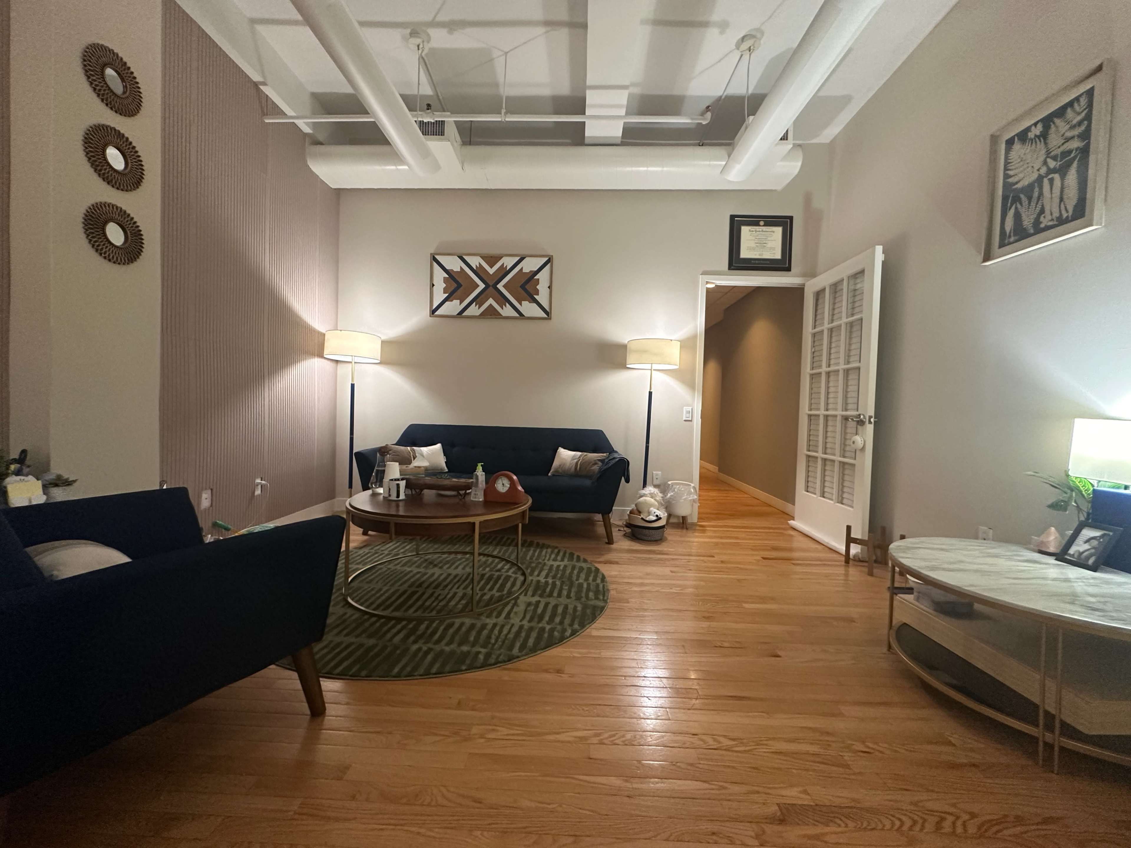 The image shows a cozy interior space featuring two navy blue sofas, a round coffee table, and a green area rug, with soft lighting from floor lamps and a side table.