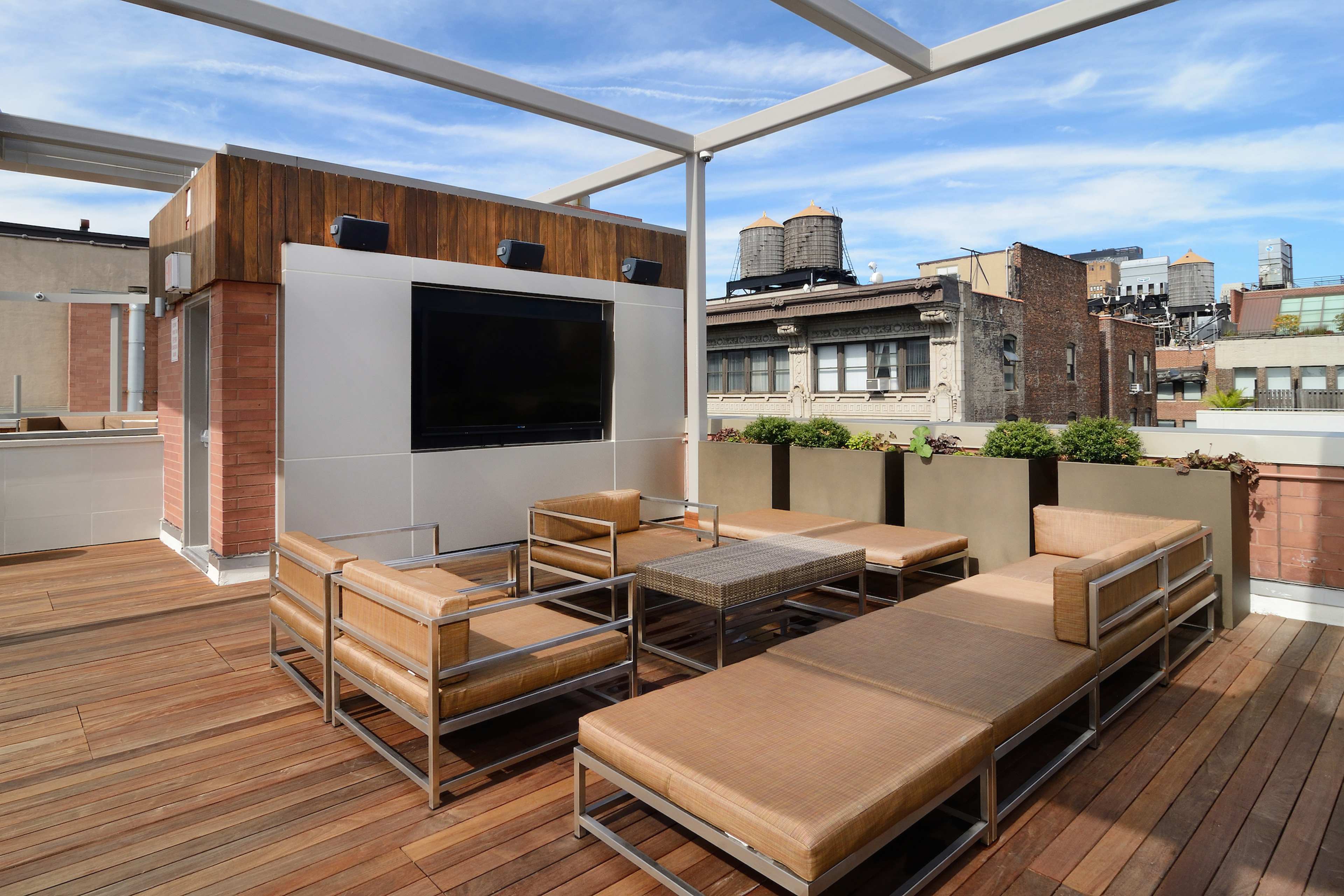 A rooftop terrace featuring modern seating arrangements and a large television mounted on a wall surrounded by city buildings.