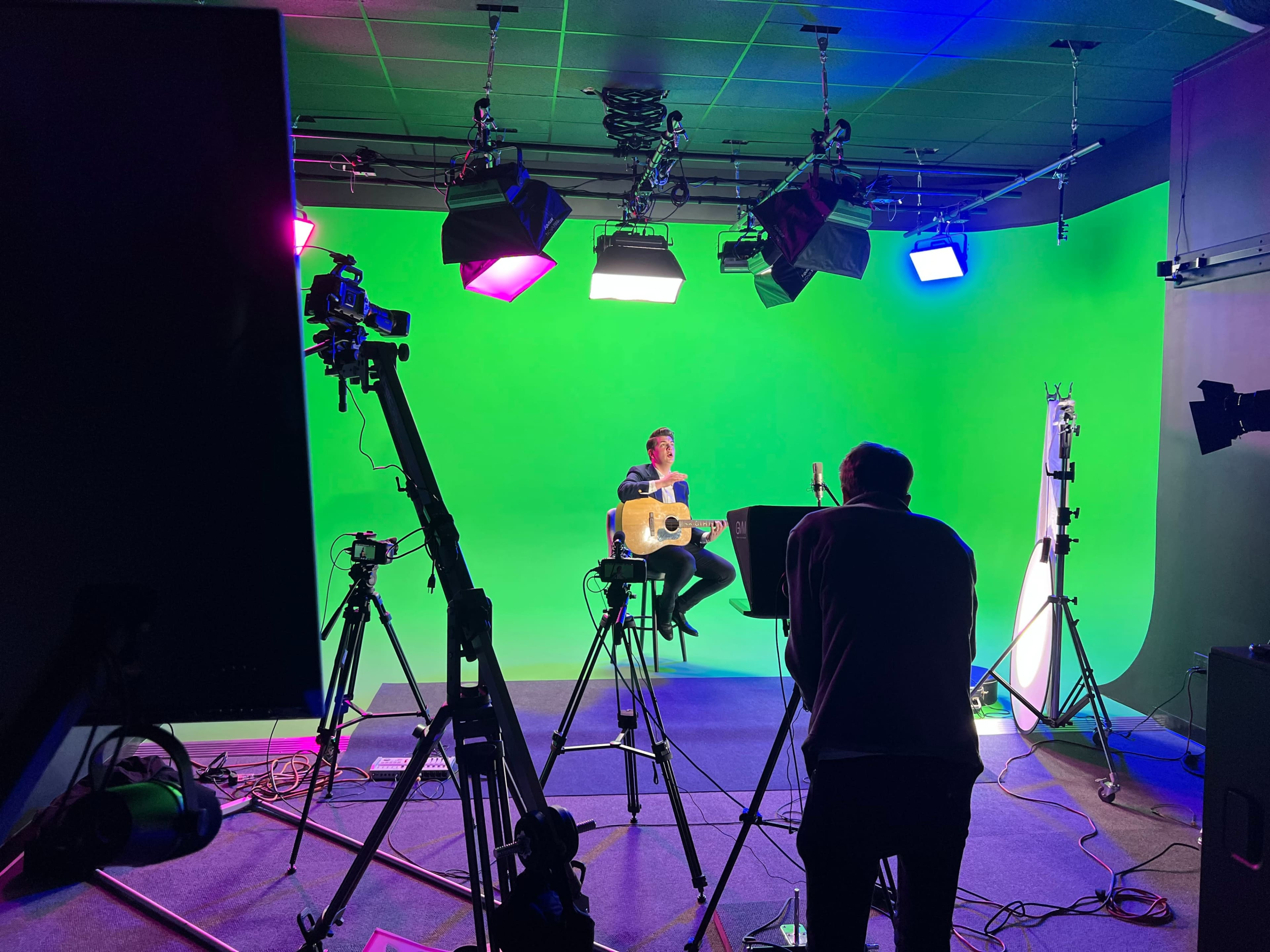 A musician sits on a stool with a guitar in front of a green screen, surrounded by multiple cameras and studio lights.