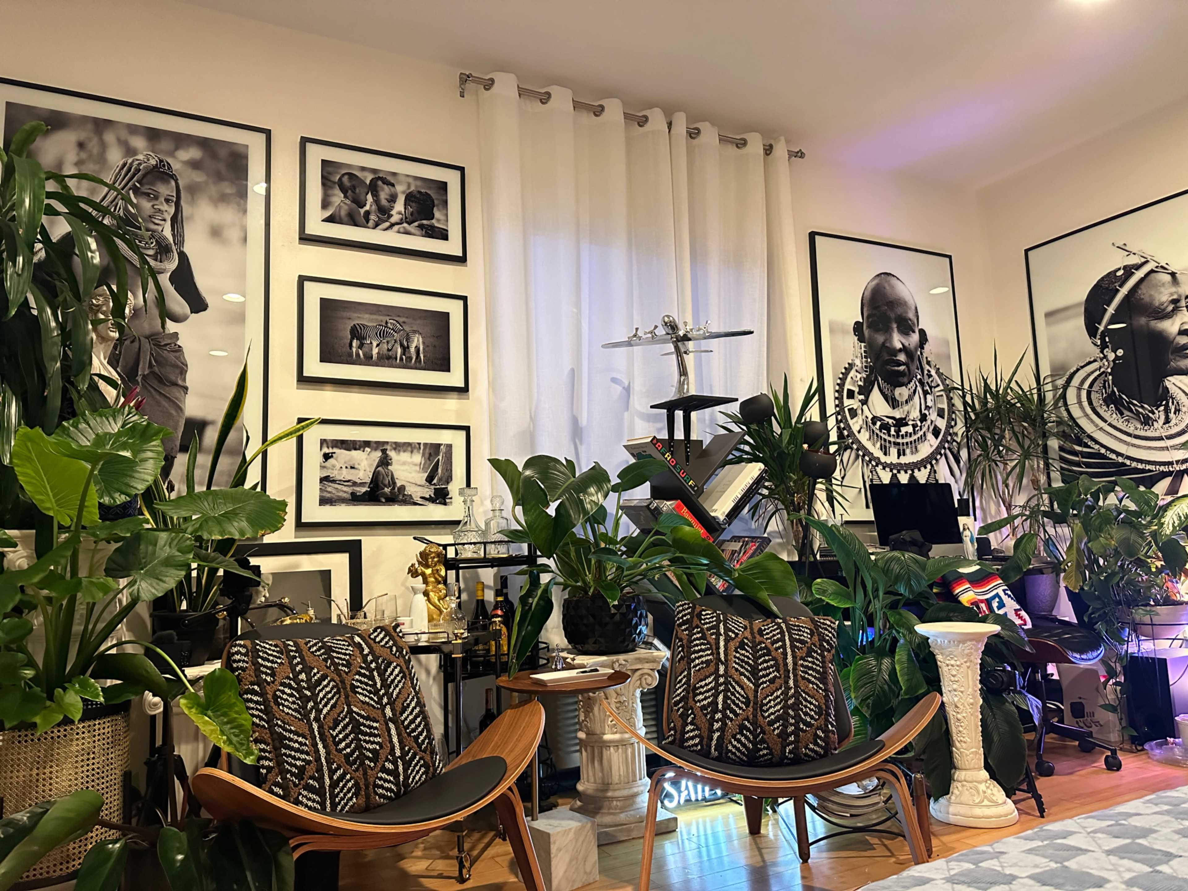 The room features two mid-century modern chairs surrounded by various indoor plants and large black-and-white photographs on the walls.