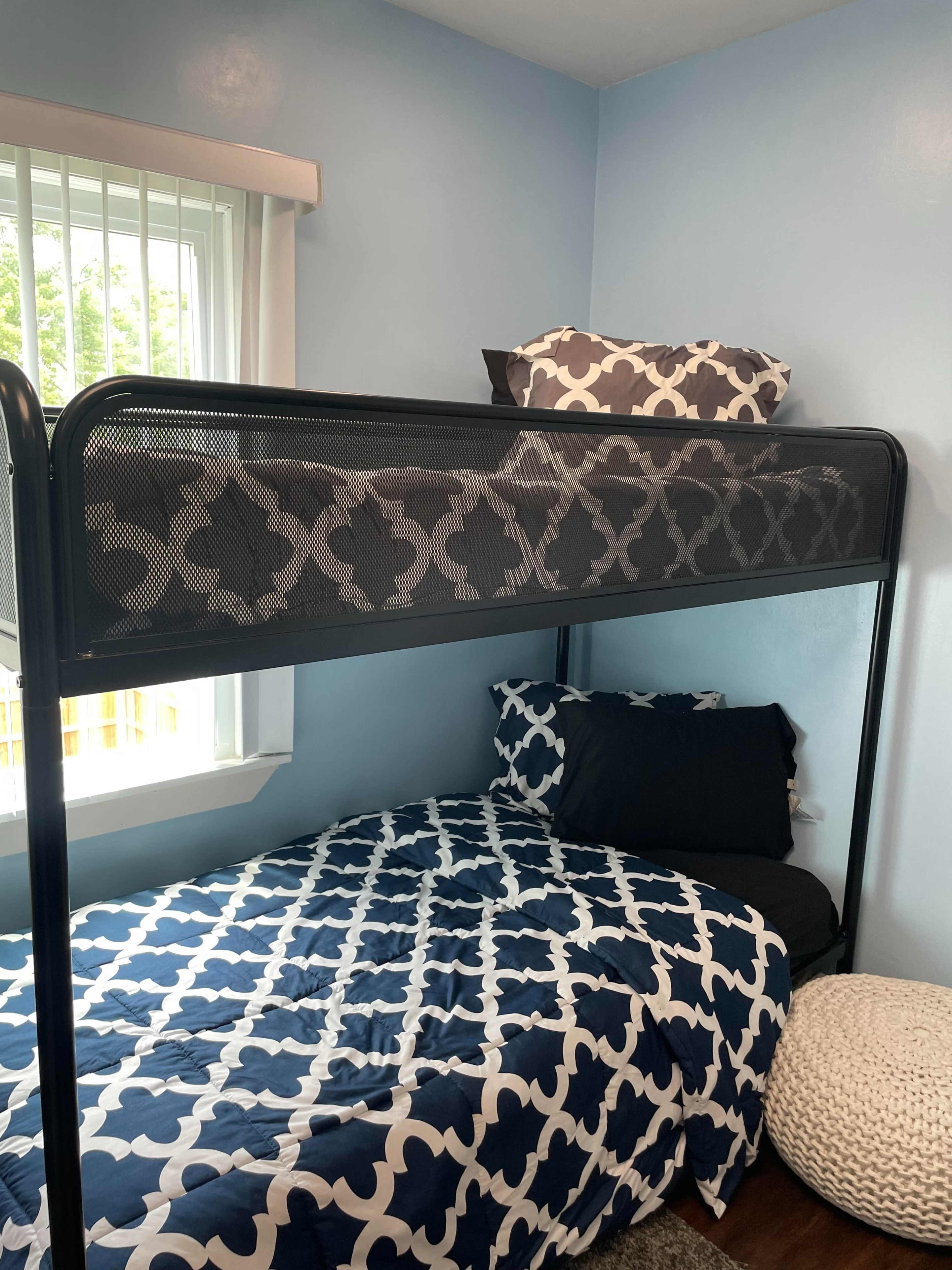 A bedroom with a lofted bed above a lower bed, both covered with a patterned blue and white bedspread, and a round pouf on the floor.