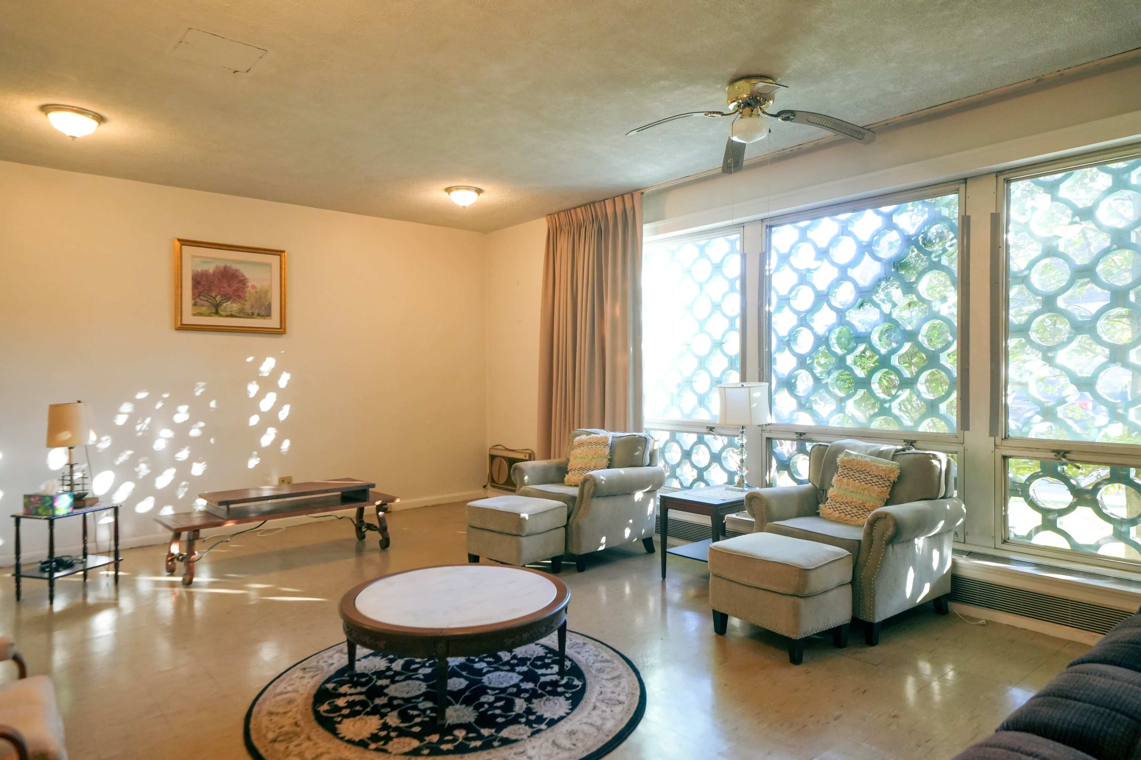 A living room features two light-colored armchairs and a circular table, illuminated by sunlight filtering through a pattern of windows.