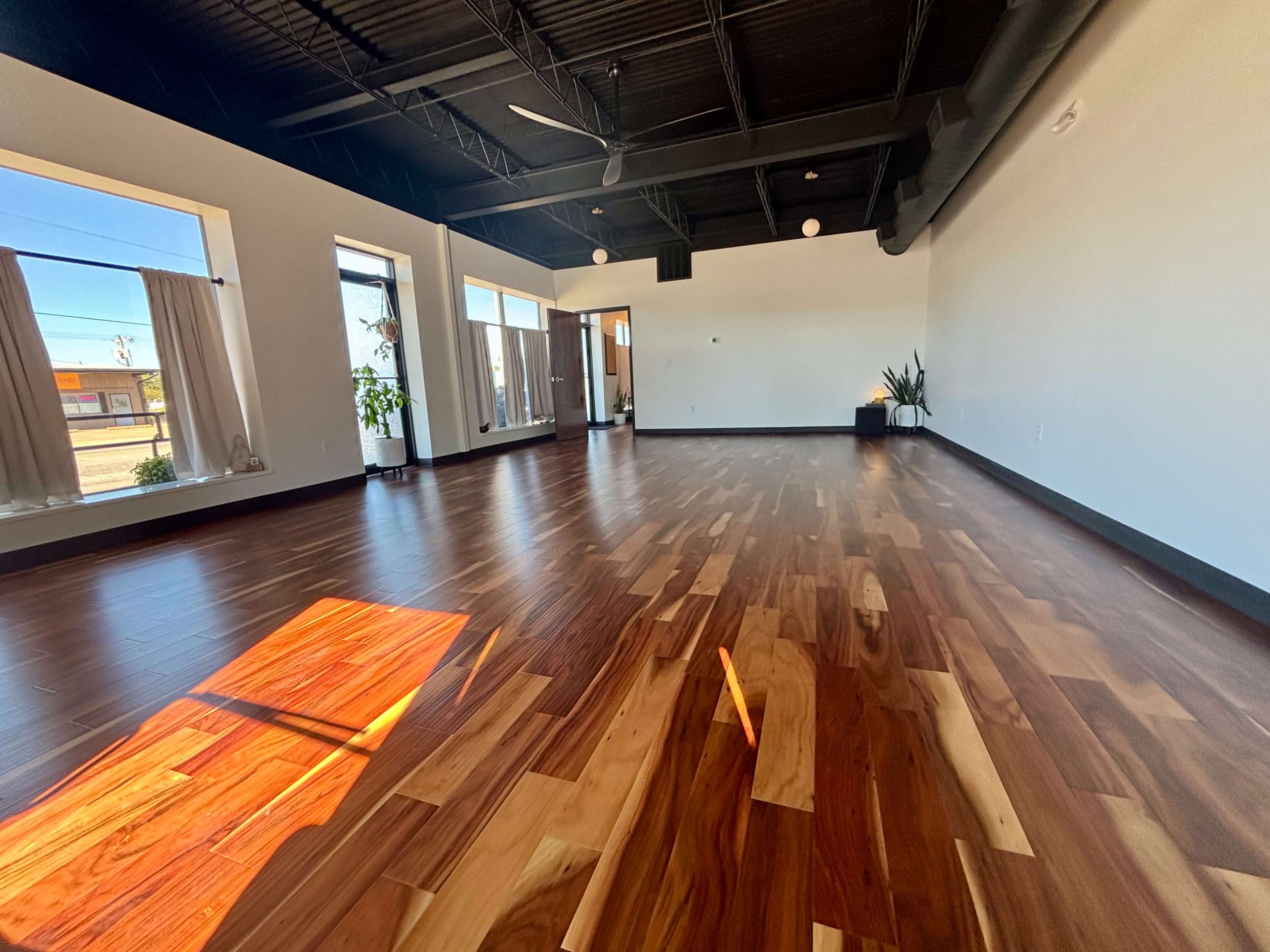 The image shows an empty room with hardwood flooring, large windows, and natural light streaming in from the left.