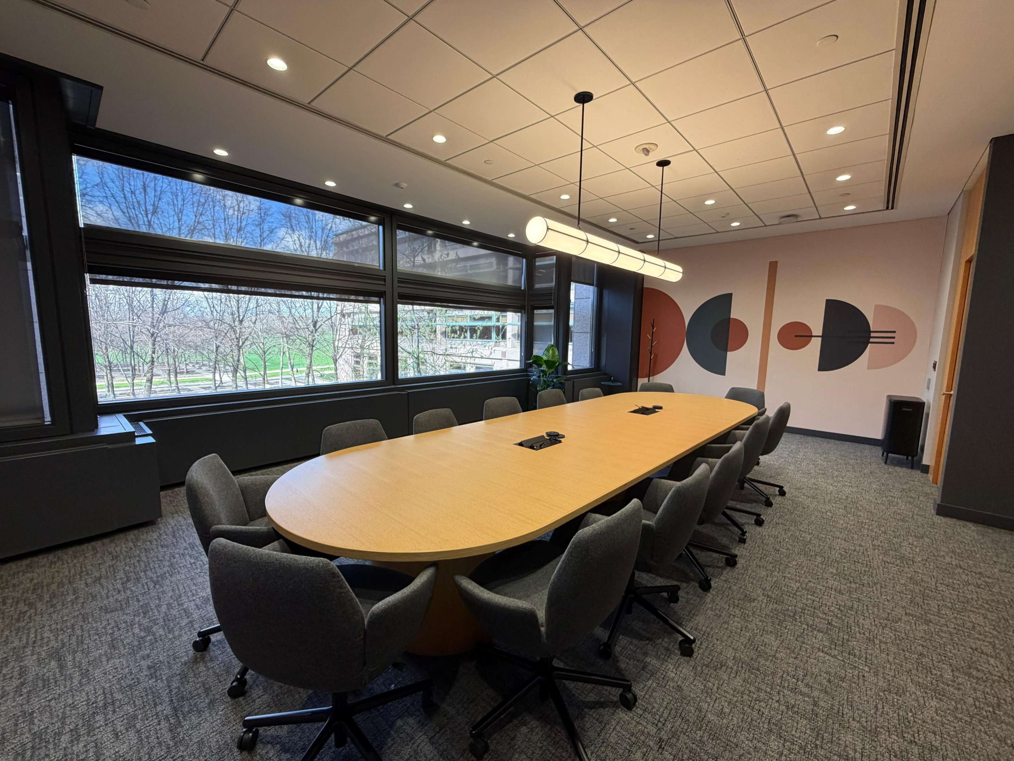 A modern conference room features a large oval table surrounded by upholstered chairs, with large windows overlooking a green landscape and abstract wall art in the background.