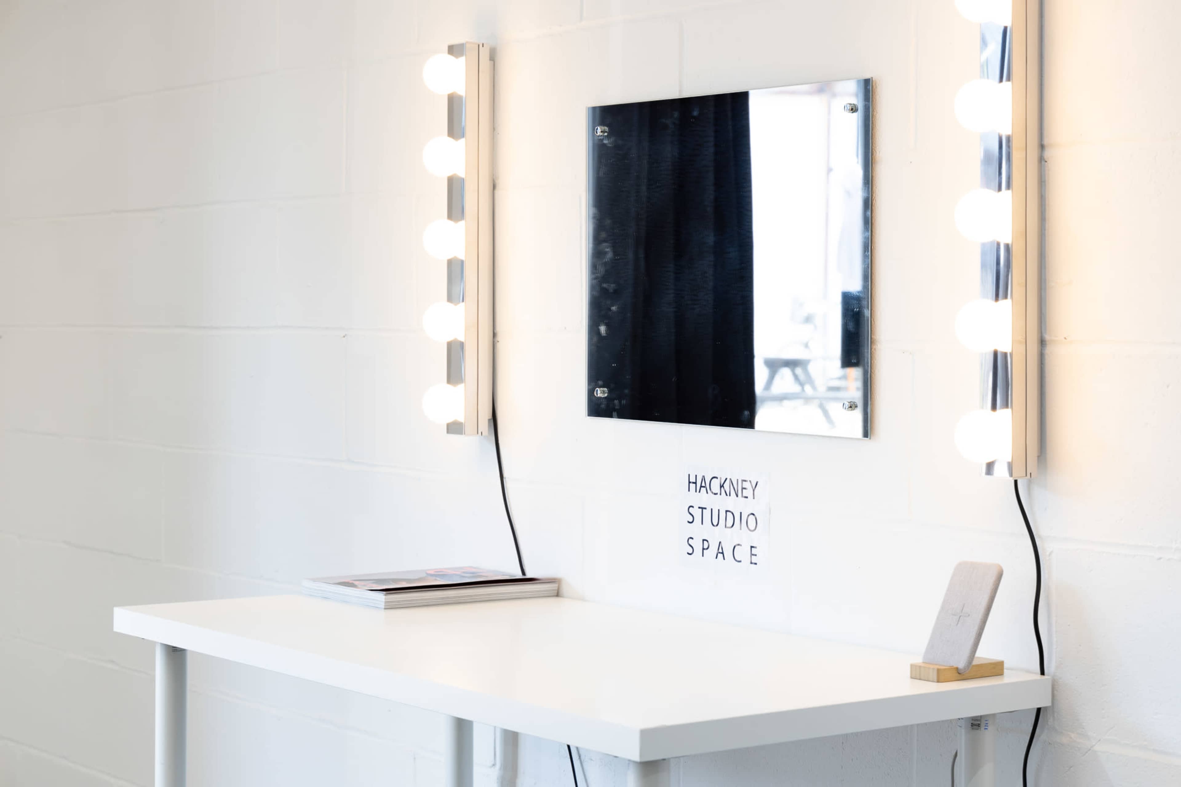 A well-lit mirror with light bulbs is mounted on a white wall above a simple table in a studio space labeled "Hackney Studio Space."