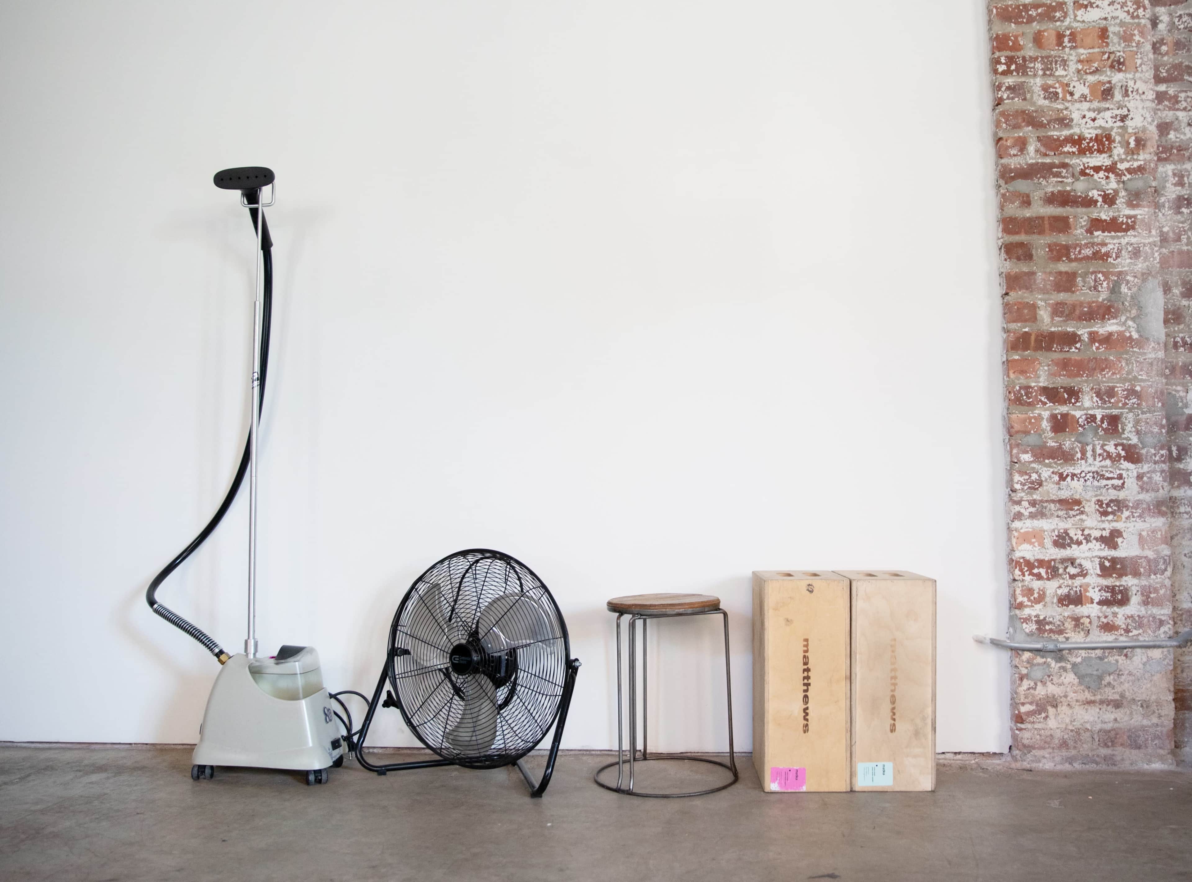 A garment steamer, a standing fan, a stool, and two stacked wooden boxes against a white wall with a brick accent.