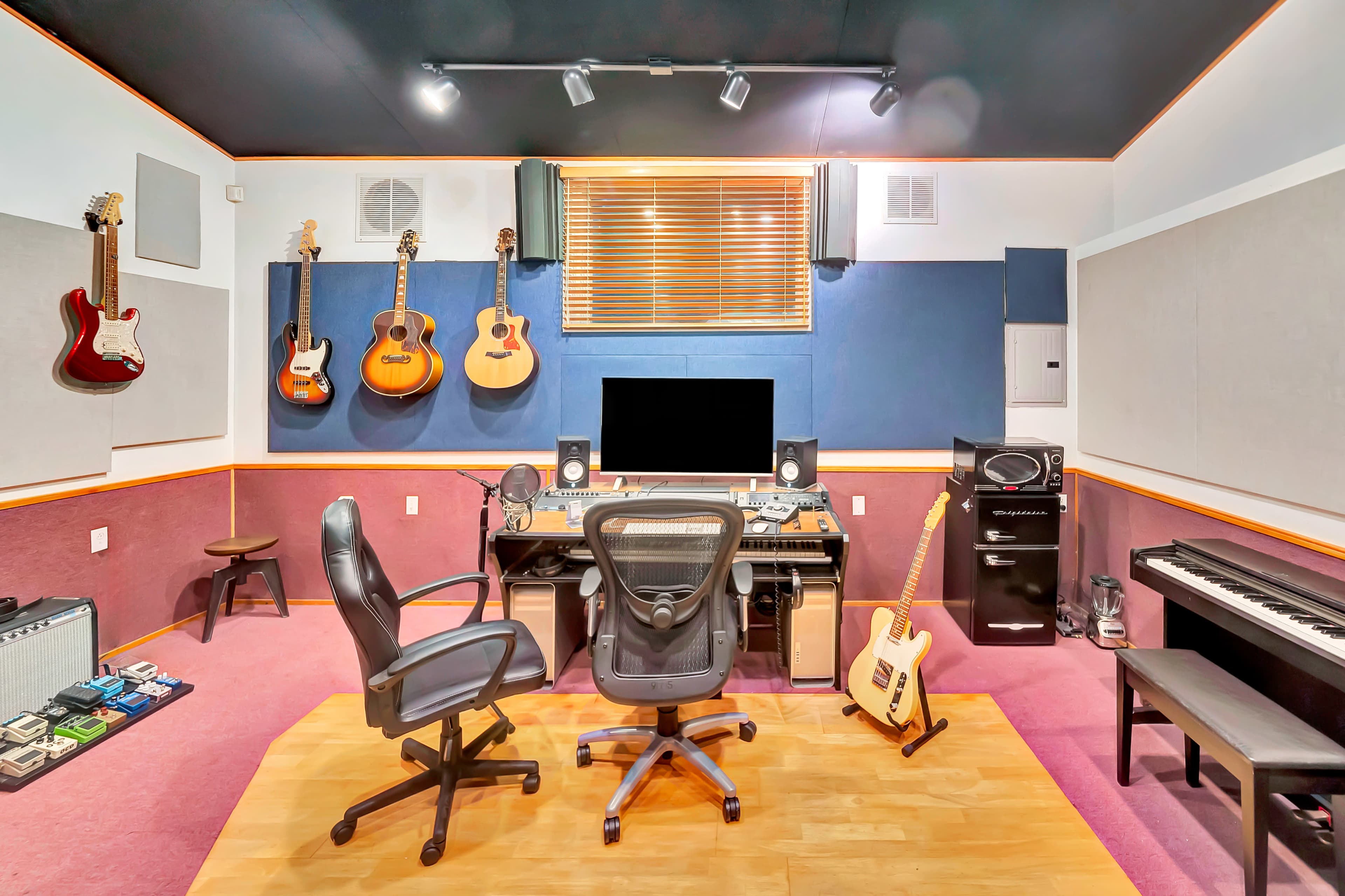 The image shows a music studio with a desk, two chairs, guitars mounted on the wall, and various musical instruments, including a keyboard and amplifiers.