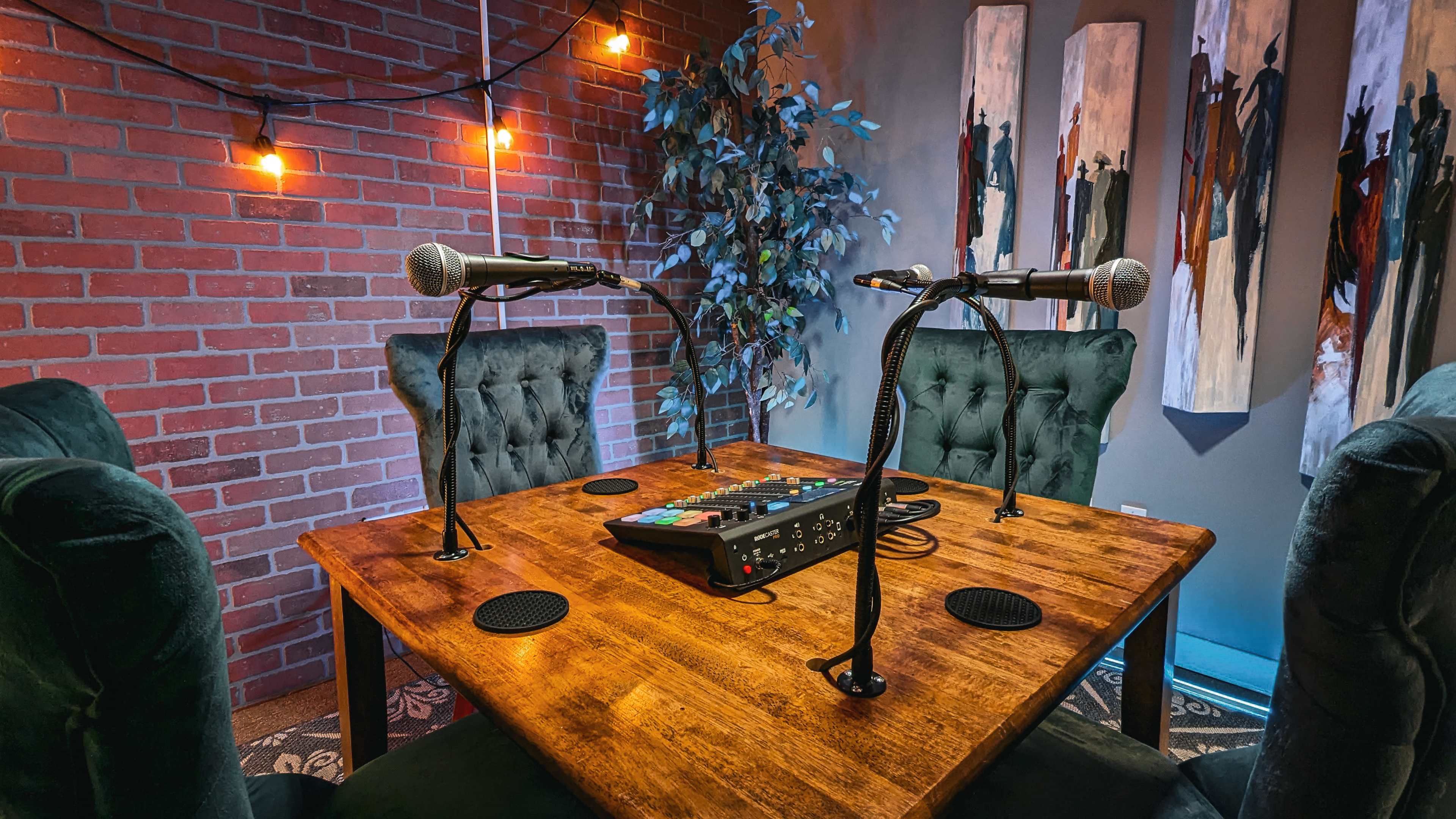 A podcast recording setup with two microphones on a wooden table, surrounded by green chairs and decorated walls featuring artwork and string lights.