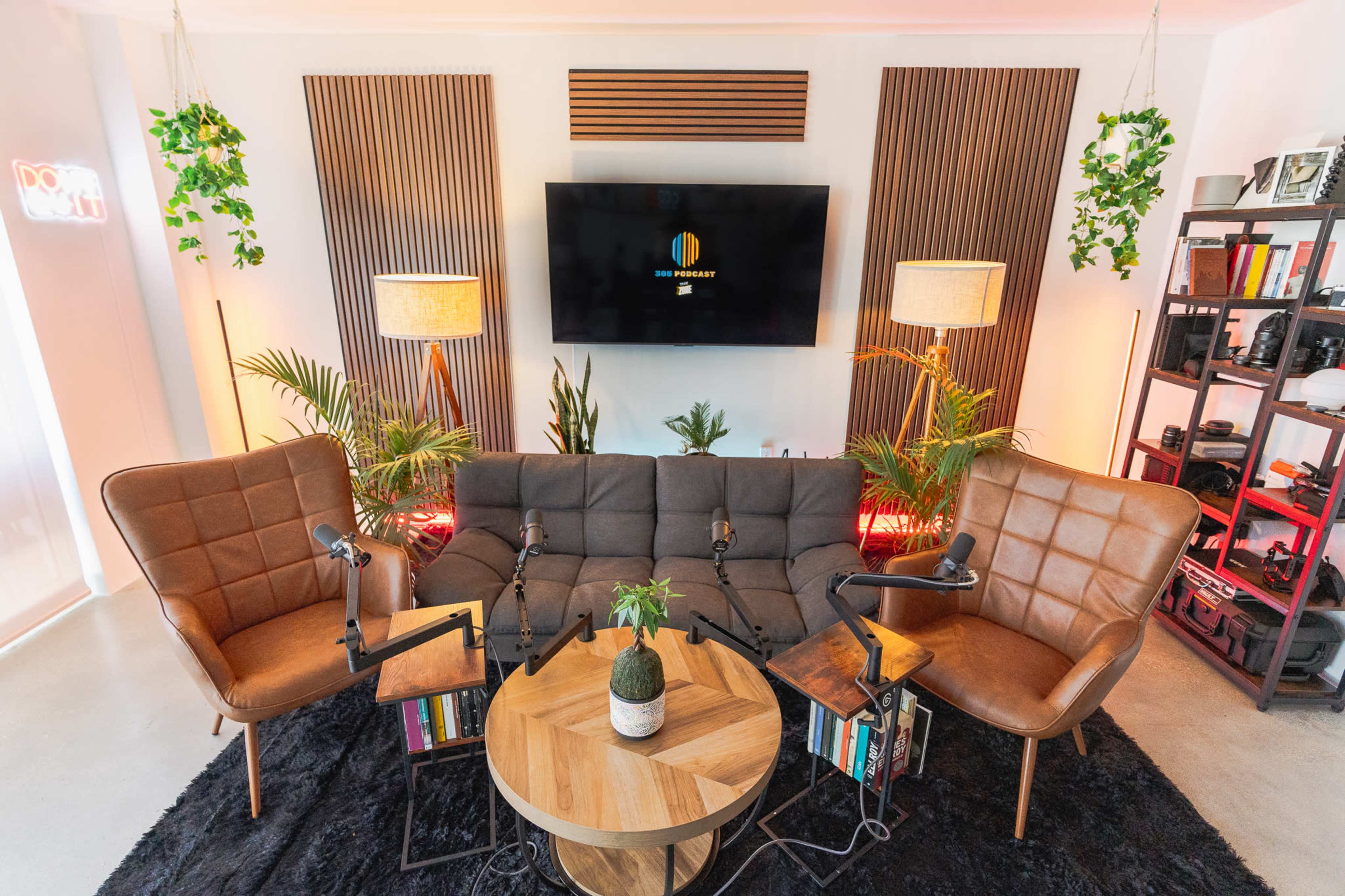 The image shows a modern podcast recording setup featuring a gray sofa and two brown armchairs around a circular wooden coffee table, with plants and lighted shelves in the background.